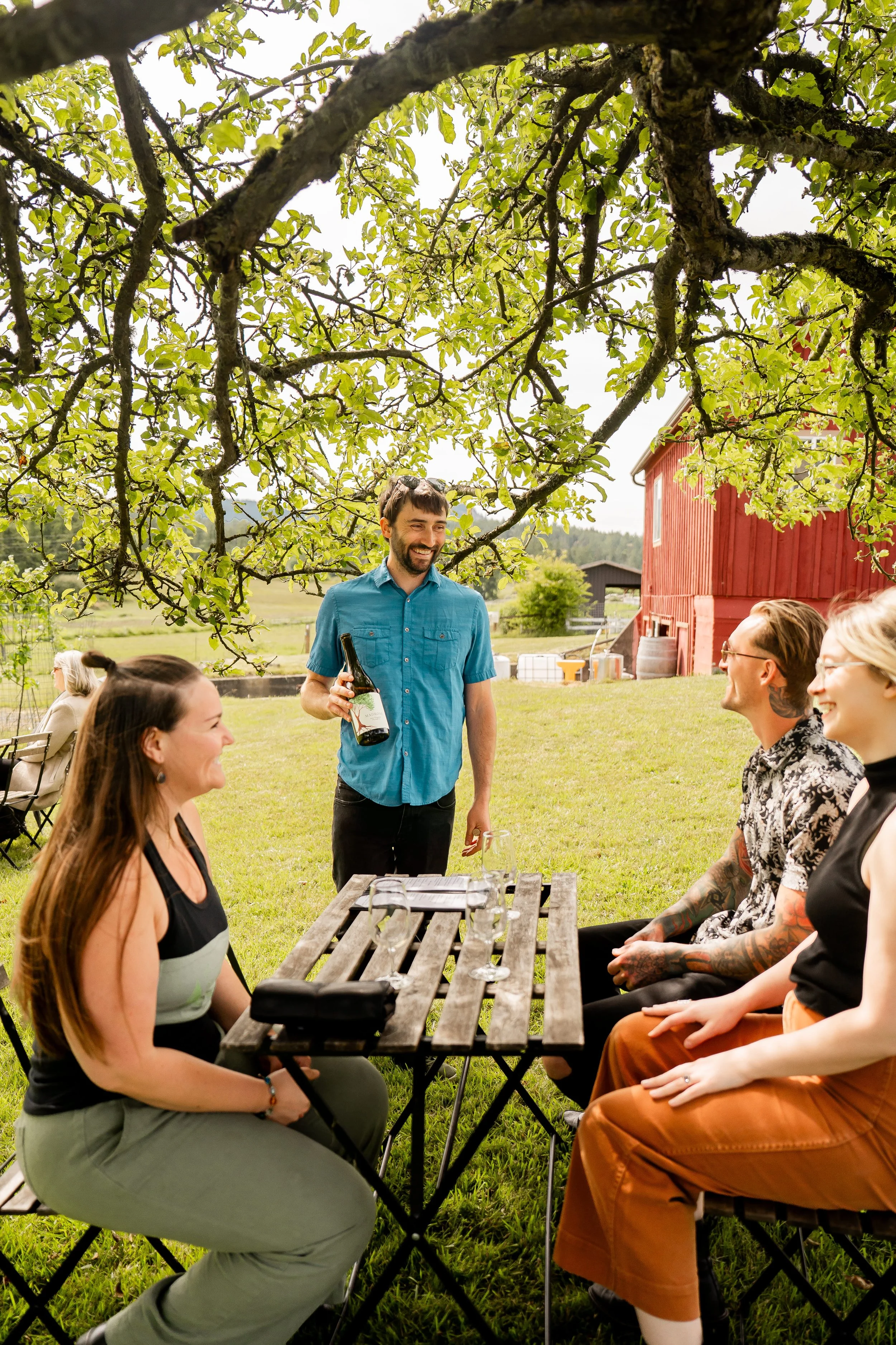Man holding wine bottle in front of wooden table with three people smiling with wine glasses on the table