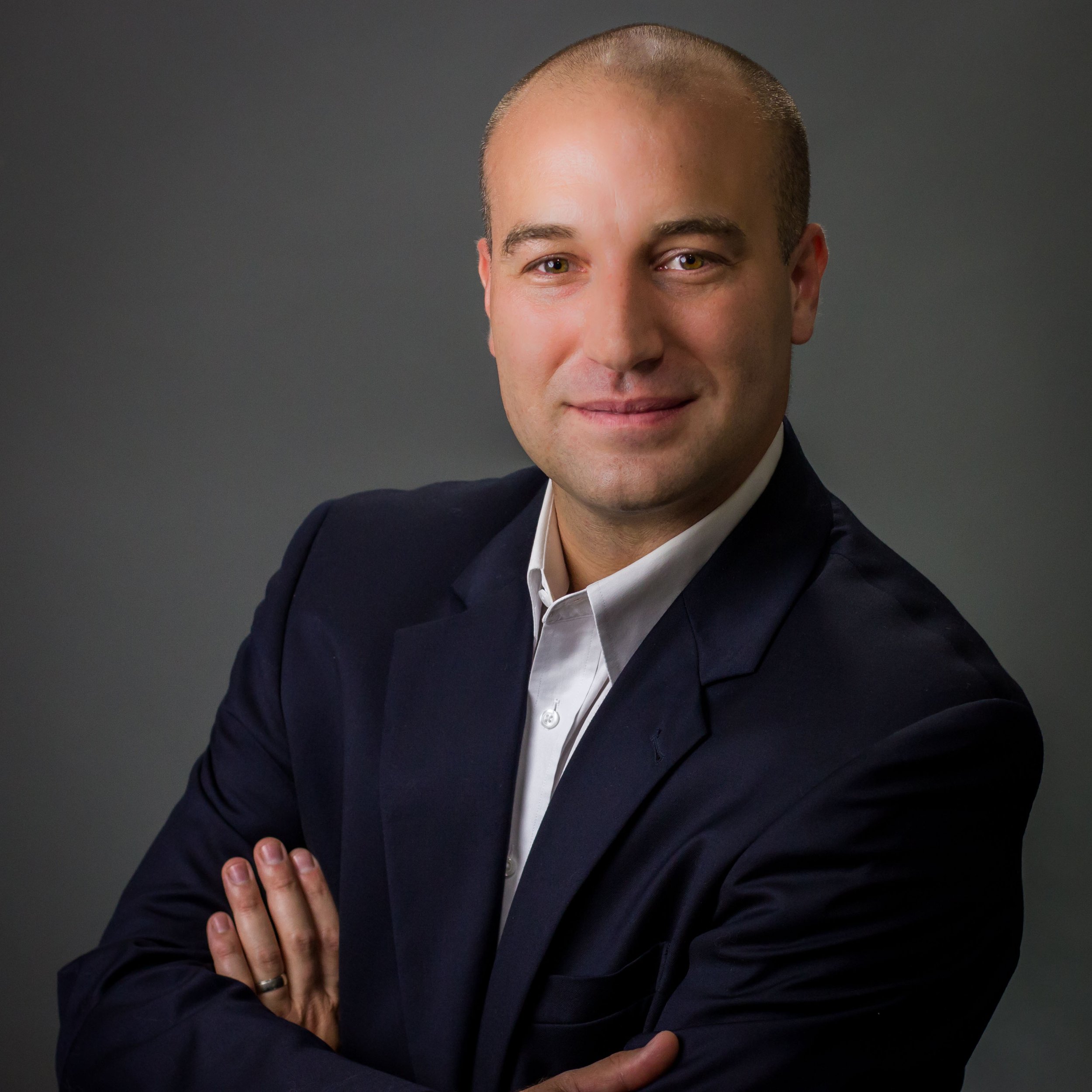 A man with a shaved head wearing a dark blazer and white shirt, arms crossed, smiling at the camera against a gray background.