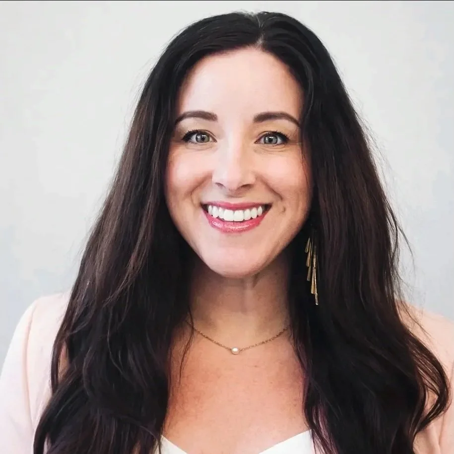 A woman with long dark hair smiling, wearing earrings, a necklace, and a light-colored top.
