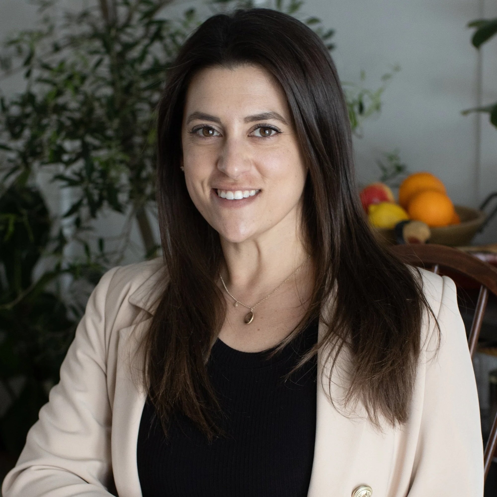 A woman with long brown hair and light skin smiling, wearing a cream blazer over a black top with a pendant necklace, sitting indoors with a bowl of fruit and green plants in the background.