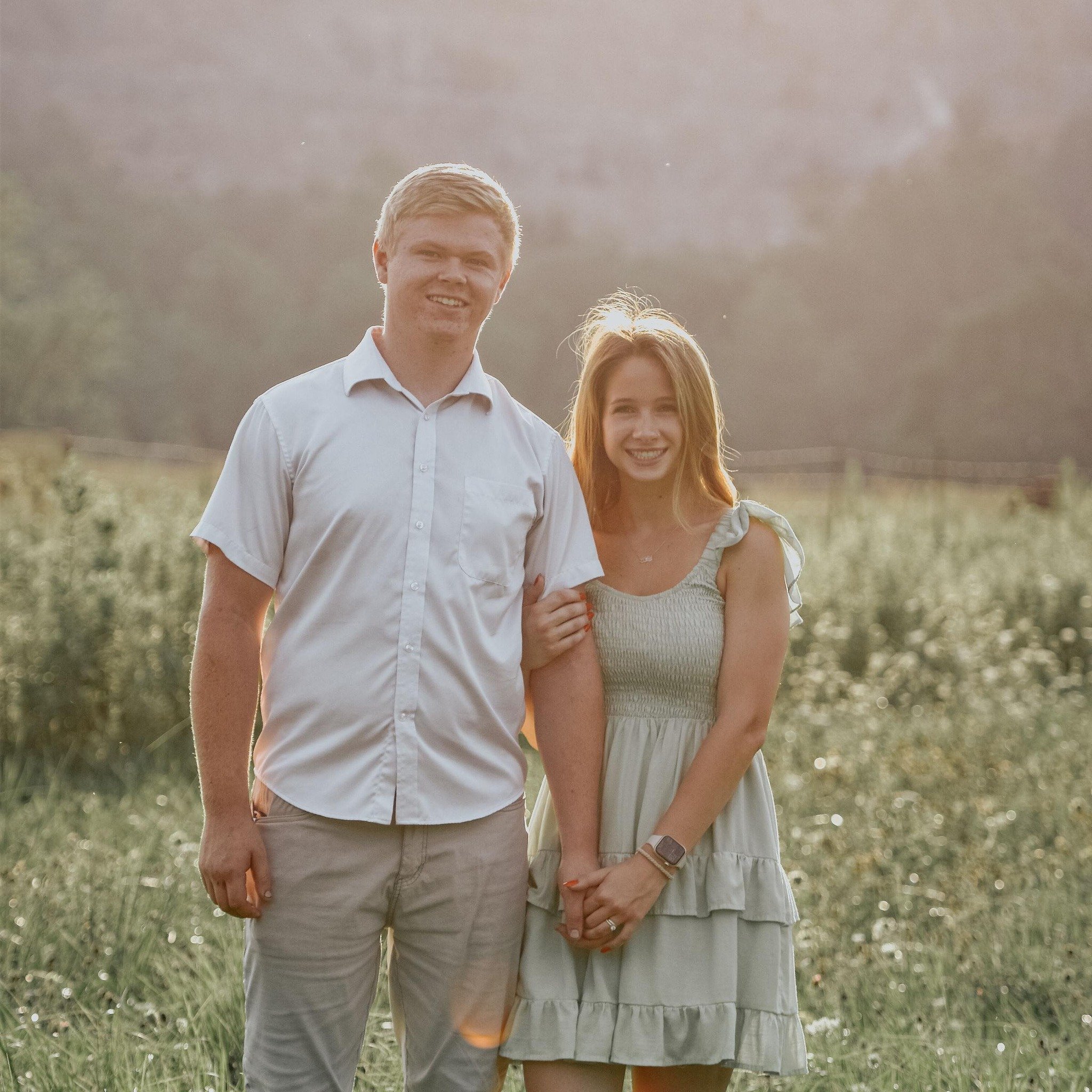 A young man and woman holding hands in a field during sunset, smiling at the camera.