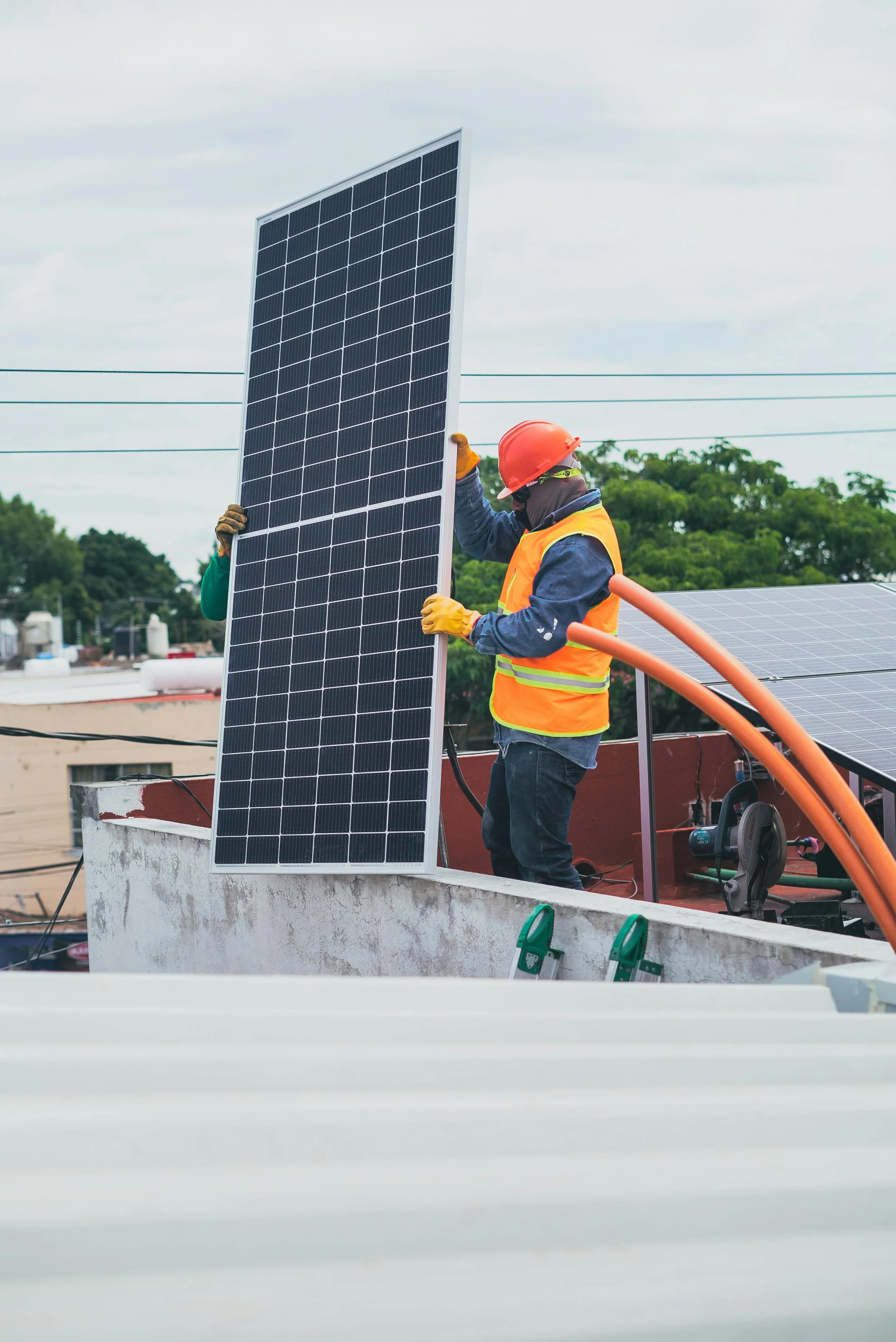 Two workers installing a solar panel on a rooftop, one wearing an orange safety vest and helmet, the other wearing a dark jacket, with green trees and electrical wires in the background.