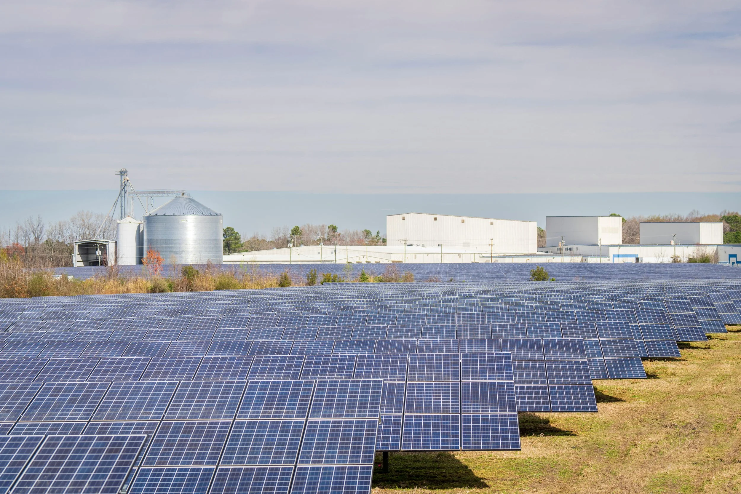 A large solar farm with numerous solar panels in the foreground and a few industrial buildings and grain silos in the background under a partly cloudy sky.
