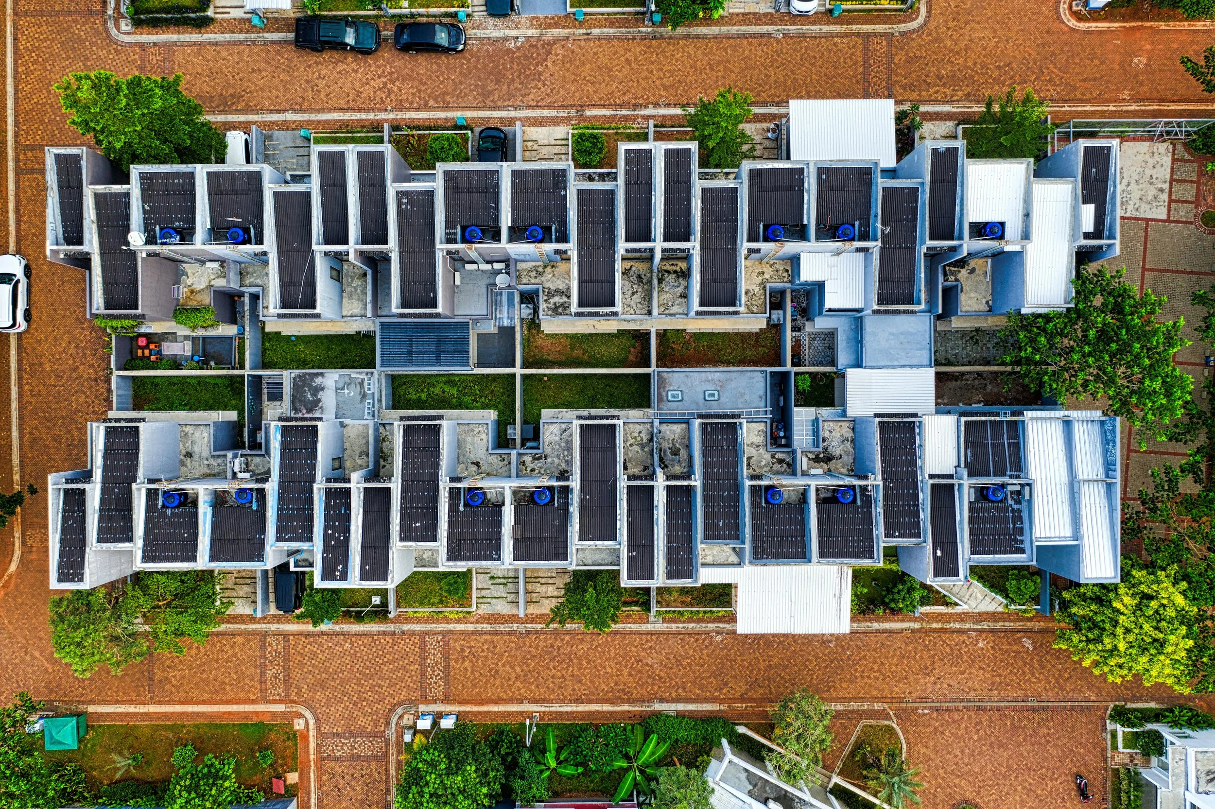 An aerial view of a modern apartment complex with multiple rooftop solar panels, surrounded by trees, brick walkways, and parked cars.