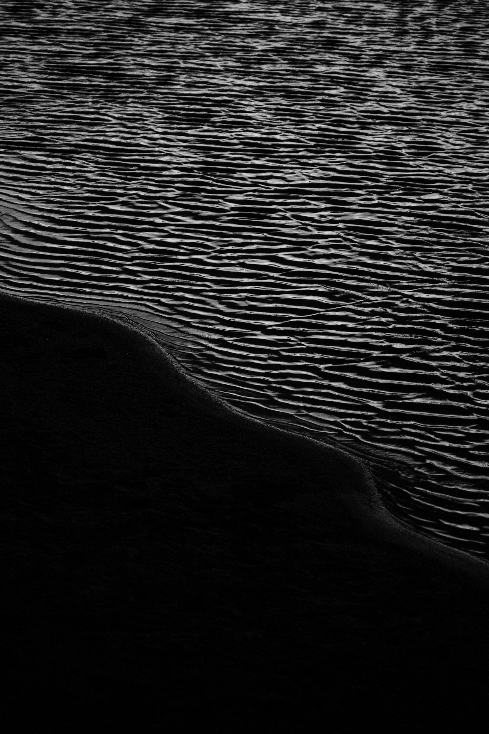Close-up of rippling water with dark sand or shoreline in the foreground, in black and white.
