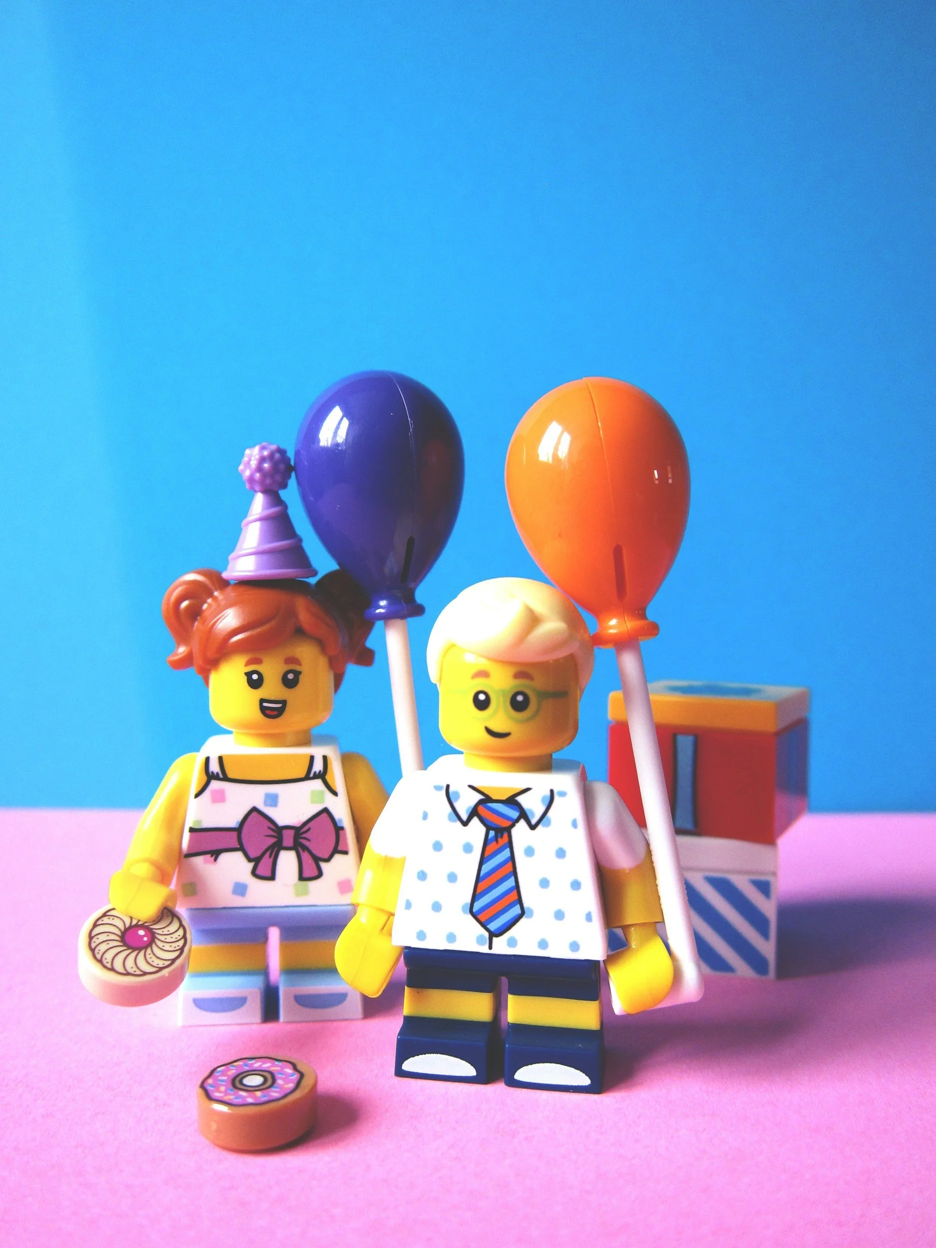 Two LEGO minifigures, a girl with red hair and a boy with blonde hair, stand against a pink and blue background, holding inflatable balloons and a doughnut, dressed in casual clothing.