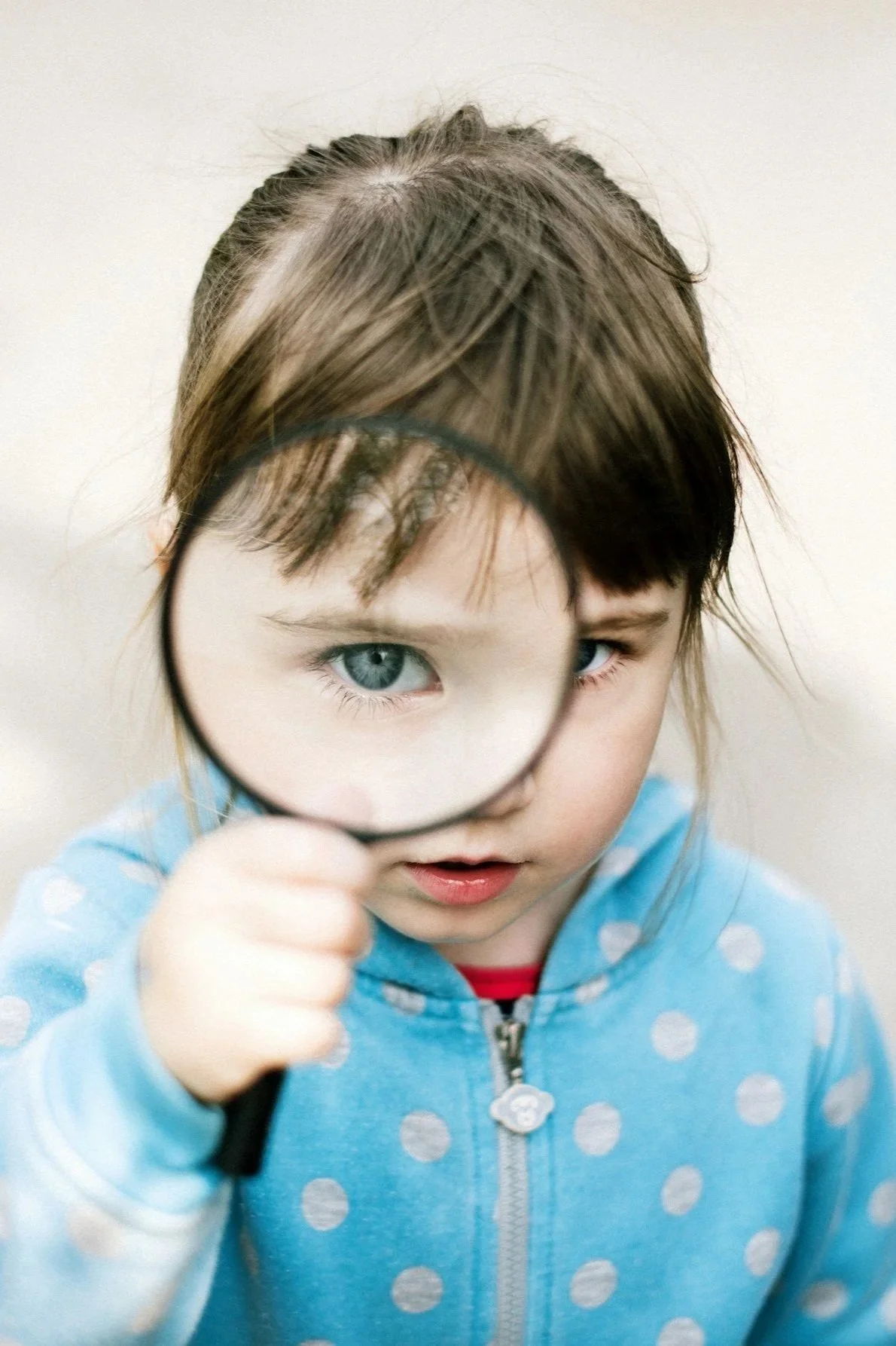 Young girl in a blue polka dot jacket holding a magnifying glass up to her face, revealing her blue eye.