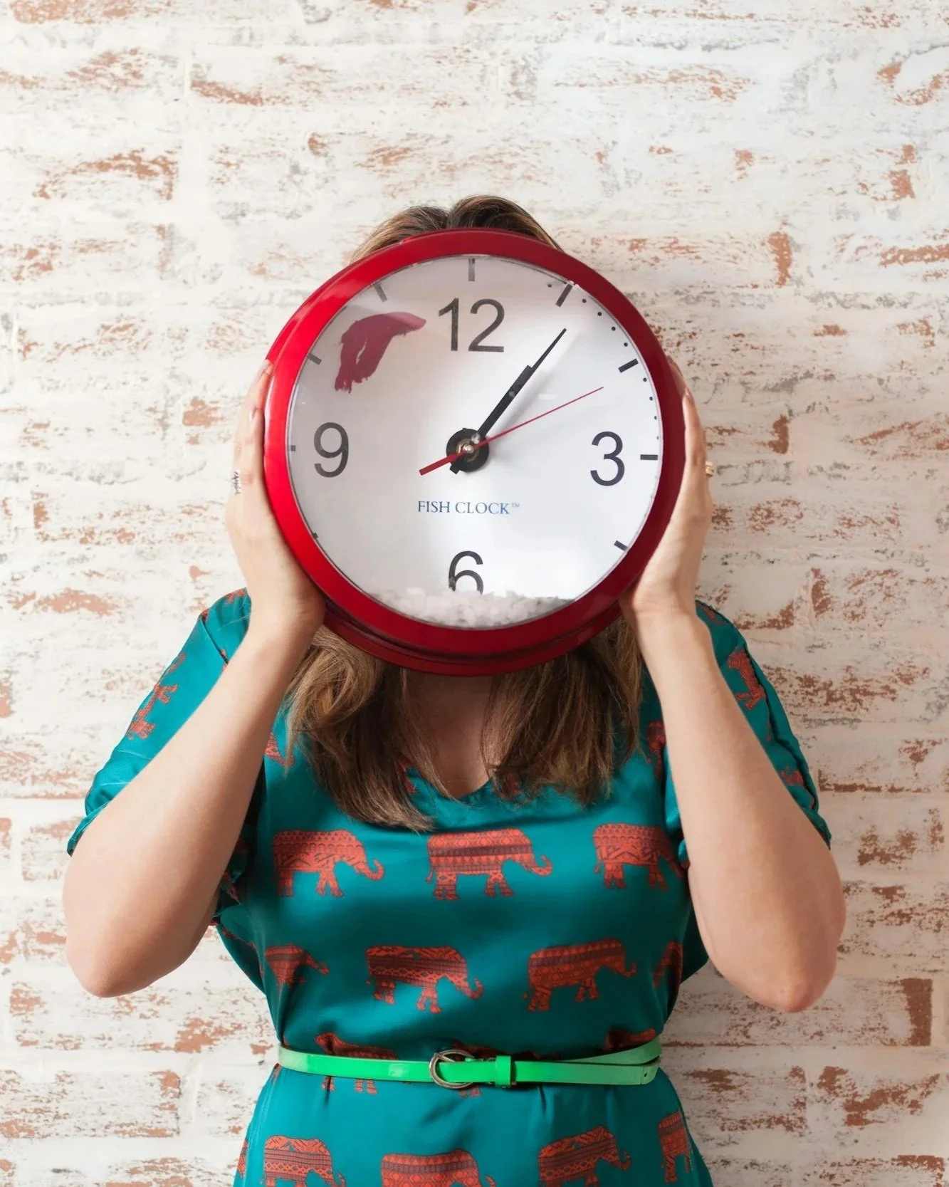 Person holding a large red Fish Clock in front of their face, standing against a white brick wall with peeling paint.