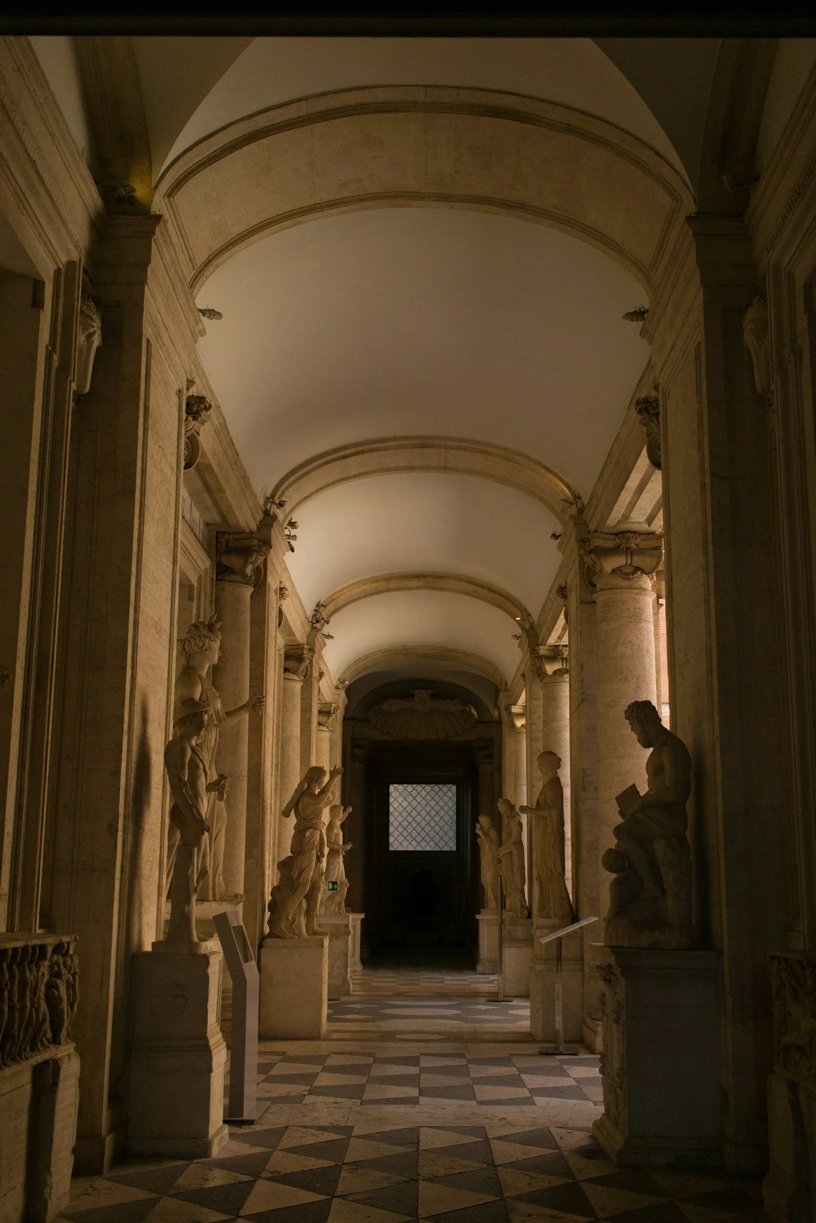 Interno di un museo con statue in marmo di figure classiche, colonnine e un soffitto arcuato, con luce soffusa e un pavimento a scacchi.