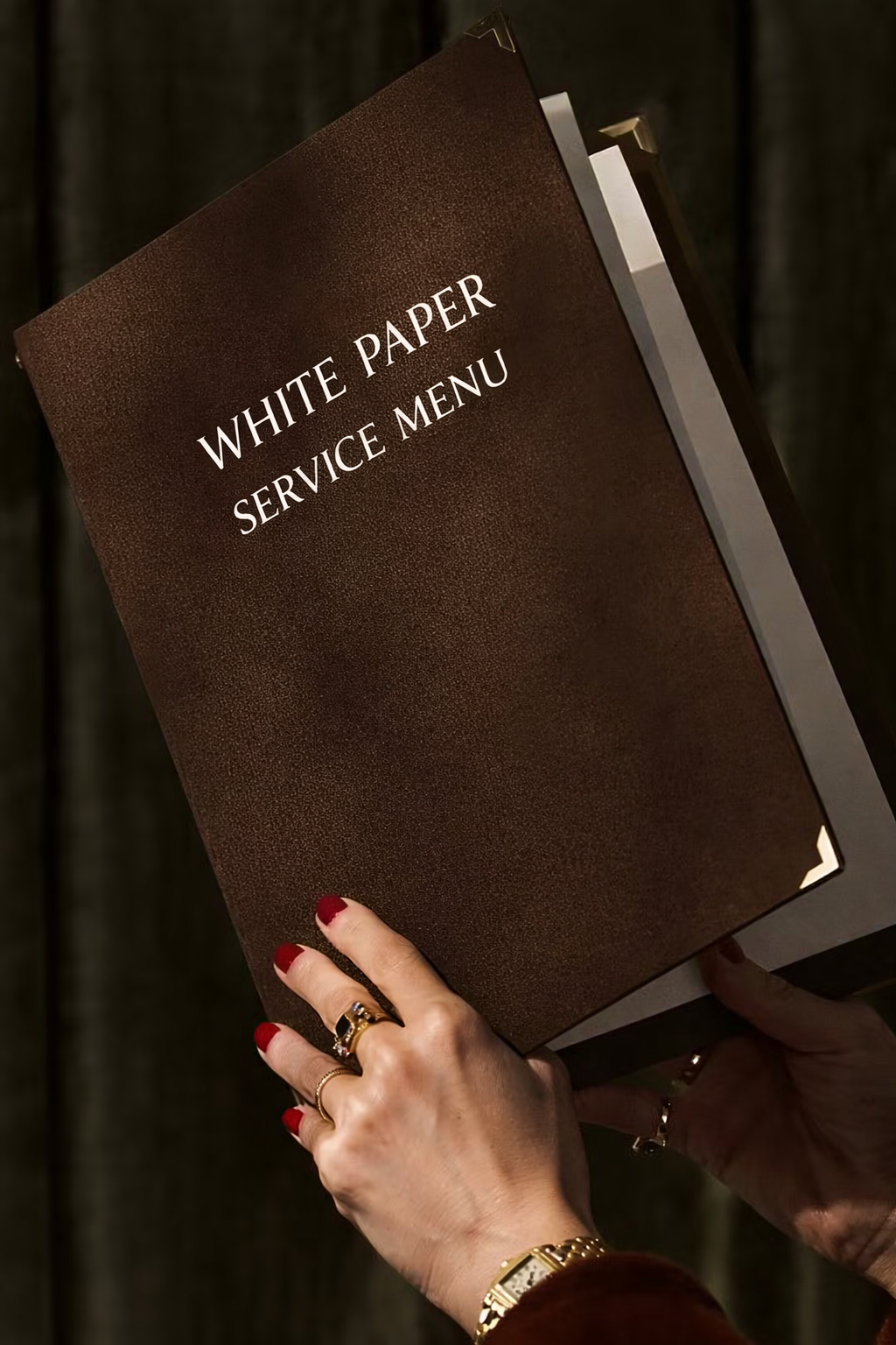 Una mano con unghie rosse tiene una serie di anelli e un orologio, e tiene in mano un menu di servizio scritto in inglese, intitolato "WHITE PAPER SERVICE MENU".