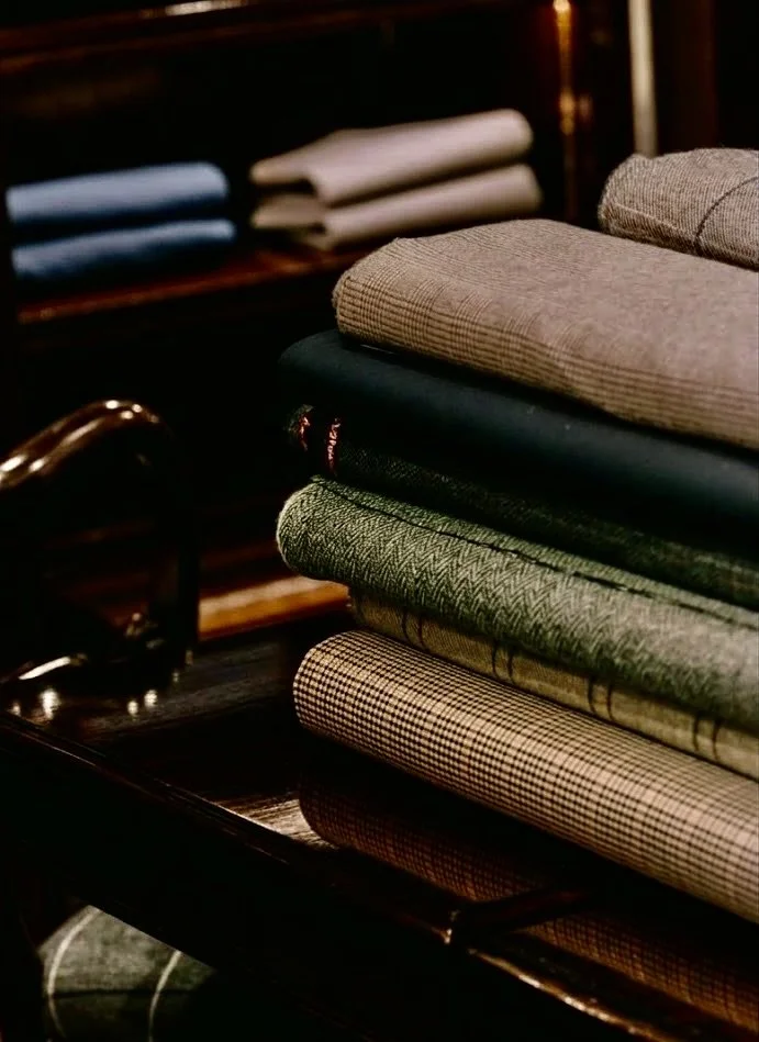 Stack di tessuti e abbigliamento da ufficio su un tavolo di legno, con sfondo di altri tessuti piegati su scaffali.