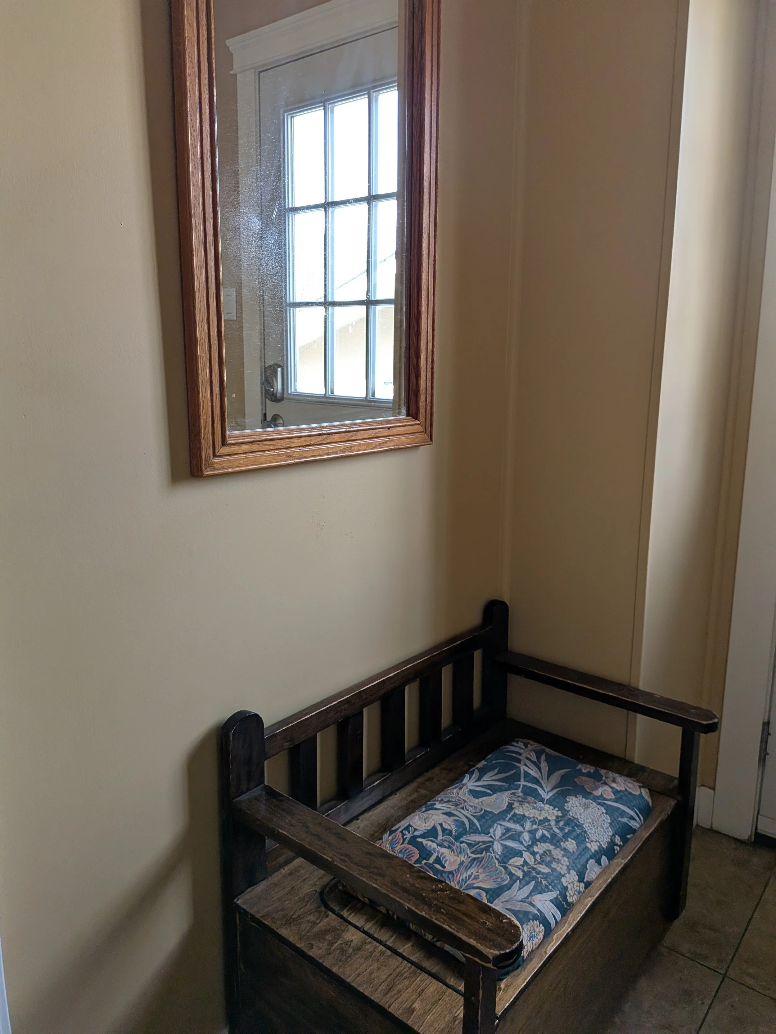 A wooden bed frame with a floral cushion, positioned against a beige wall. A mirror hanging on the wall reflects a window with divided panes and a door handle.