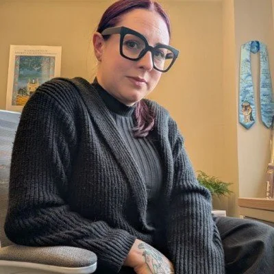Woman with red hair, black glasses, wearing a black sweater, seated indoors, with a framed picture and a blue ribbon hanging on the wall behind her.
