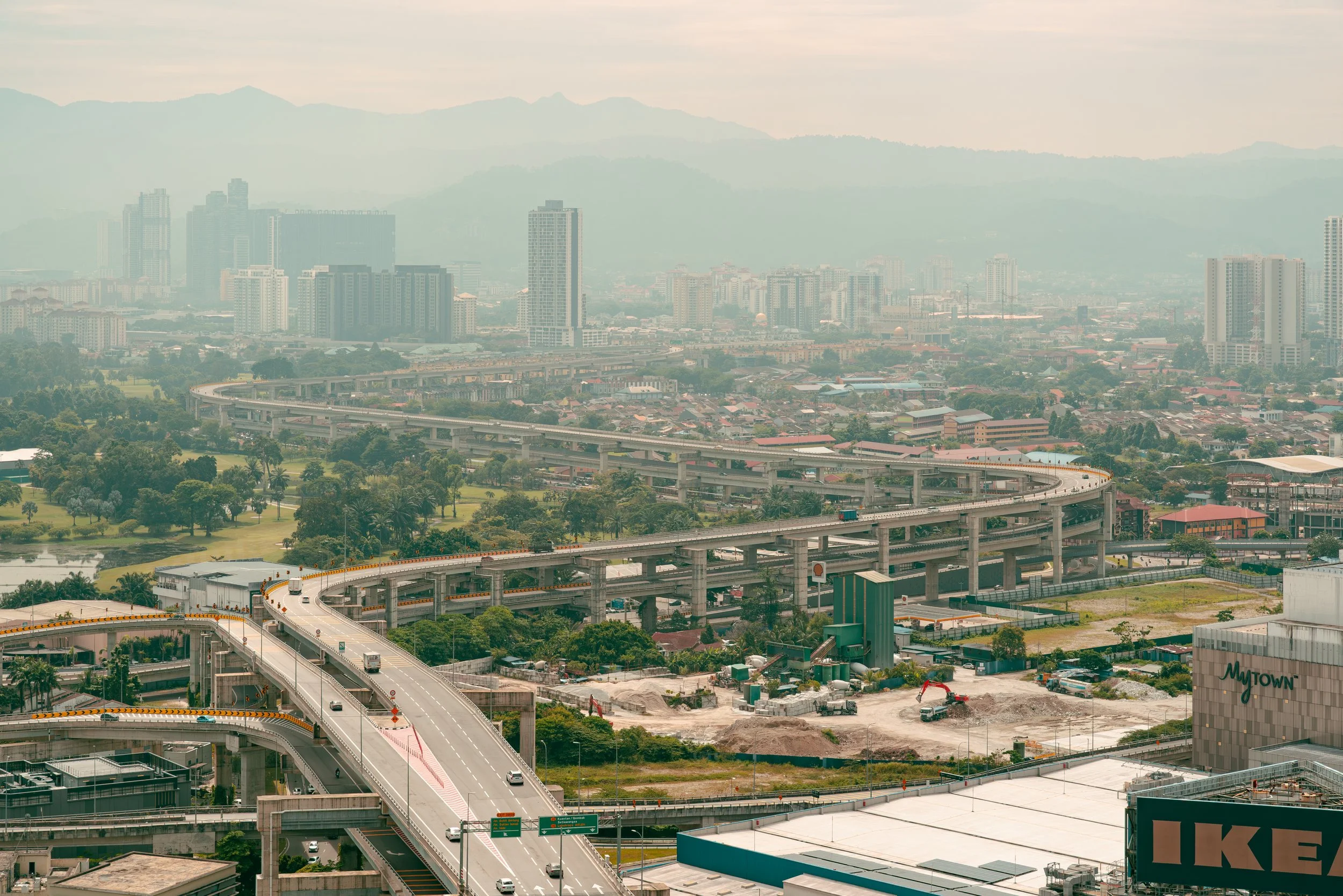 Cityscape with elevated highways, high-rise buildings, trees, and mountains in the background.