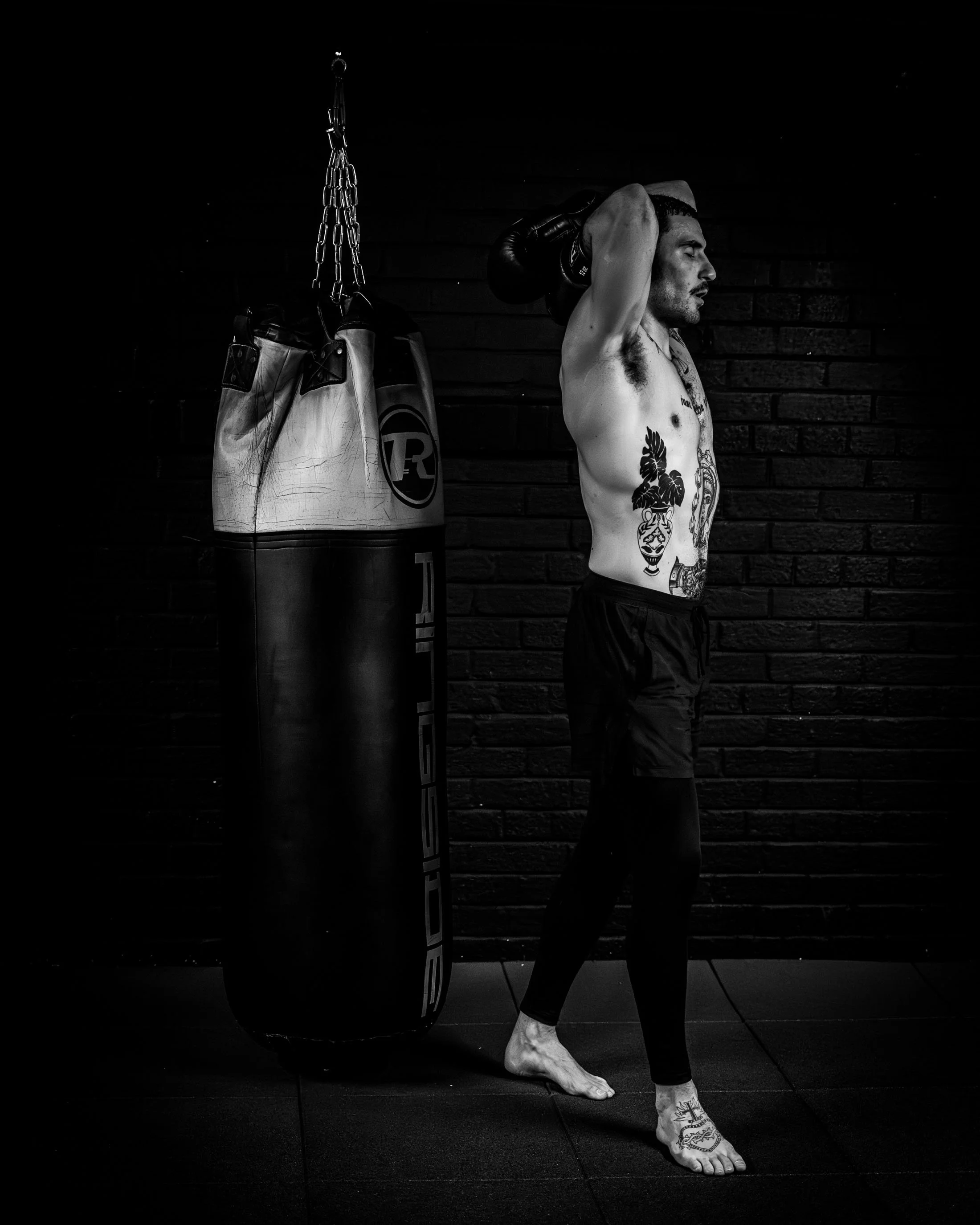 A shirtless man with tattoos stands in a gym next to a hanging punching bag, holding boxing gloves on his head, with a brick wall in the background.