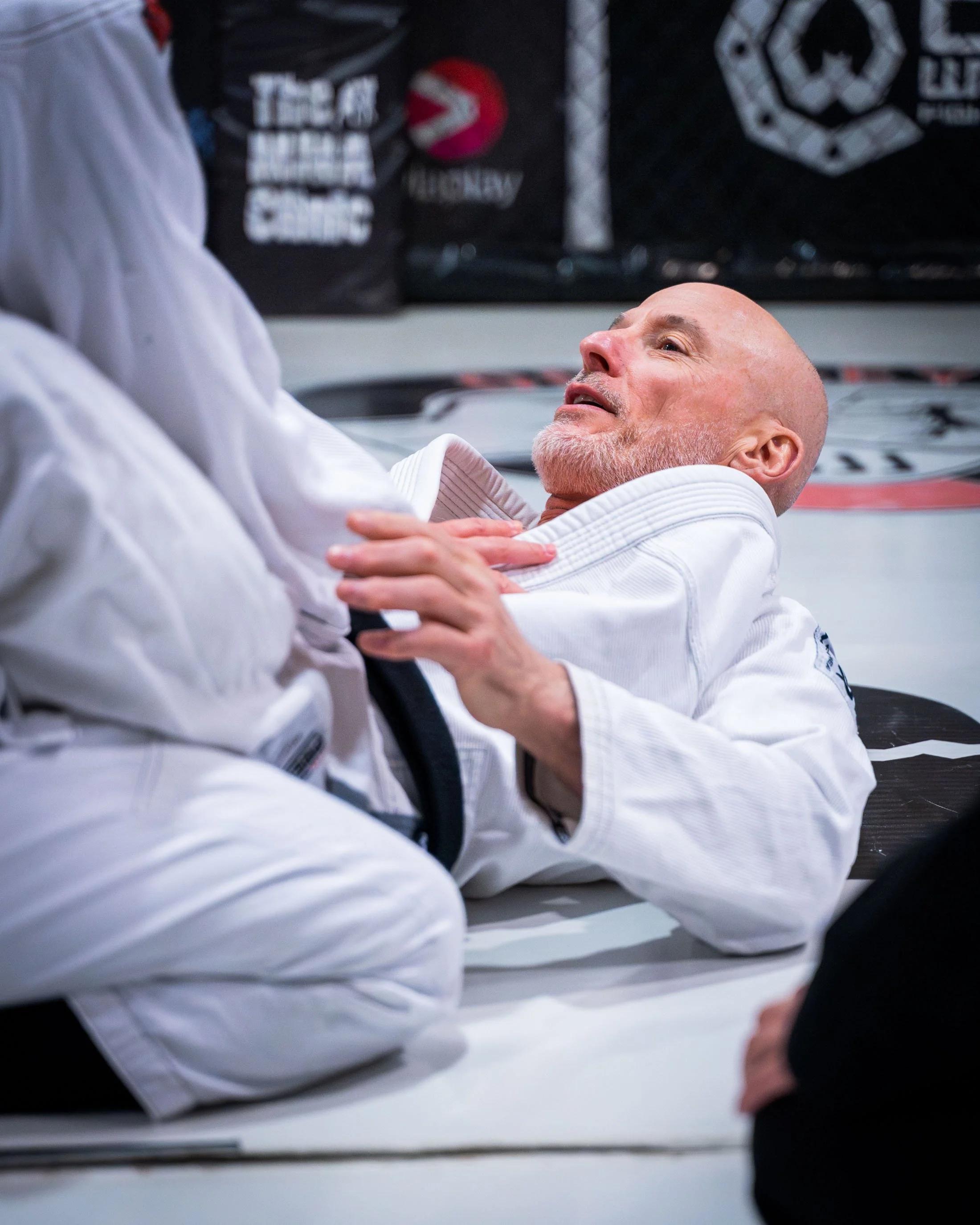 A man in a white Brazilian Jiu-Jitsu gi lying on the mat, holding onto his opponent's collar during a grappling match in a gym.