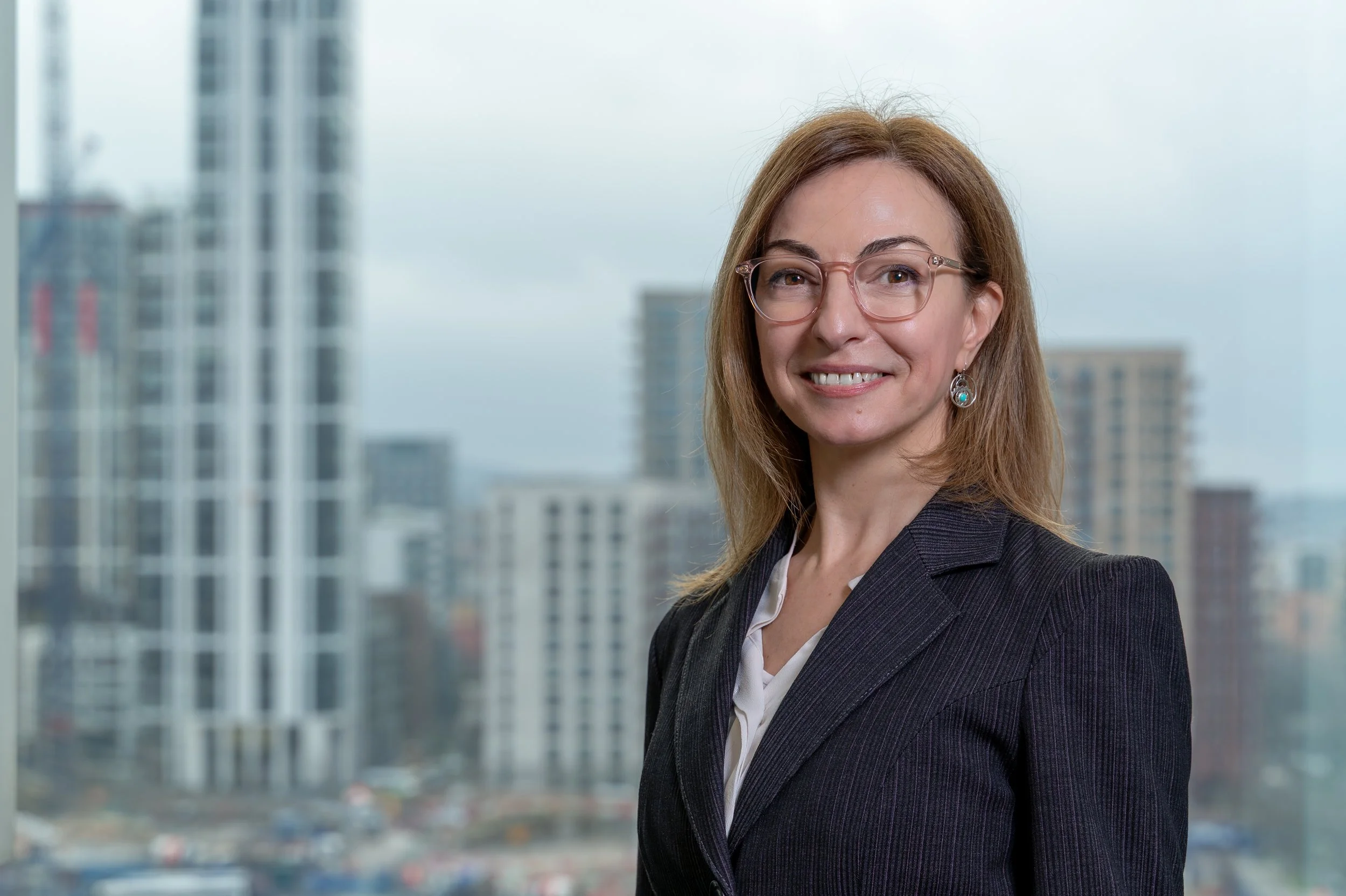 Professional woman with glasses and earrings smiling in an office with city buildings outside the window.