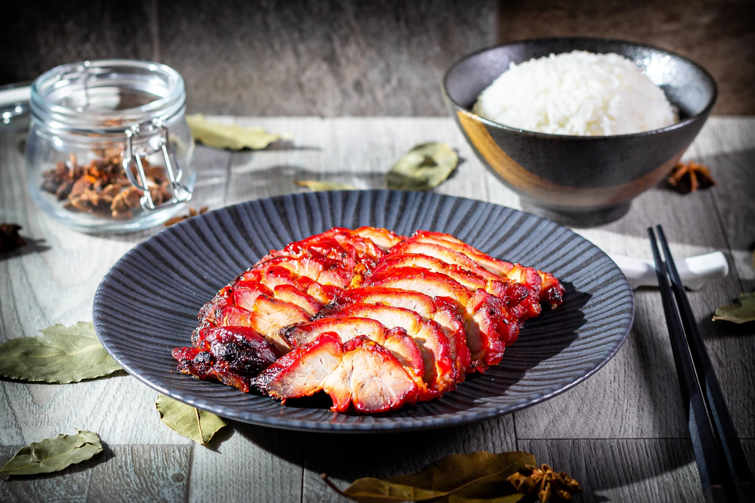 Sliced roasted meat on a dark plate with rice in a black bowl behind it, on a wooden table with bay leaves and a glass jar of spices nearby.