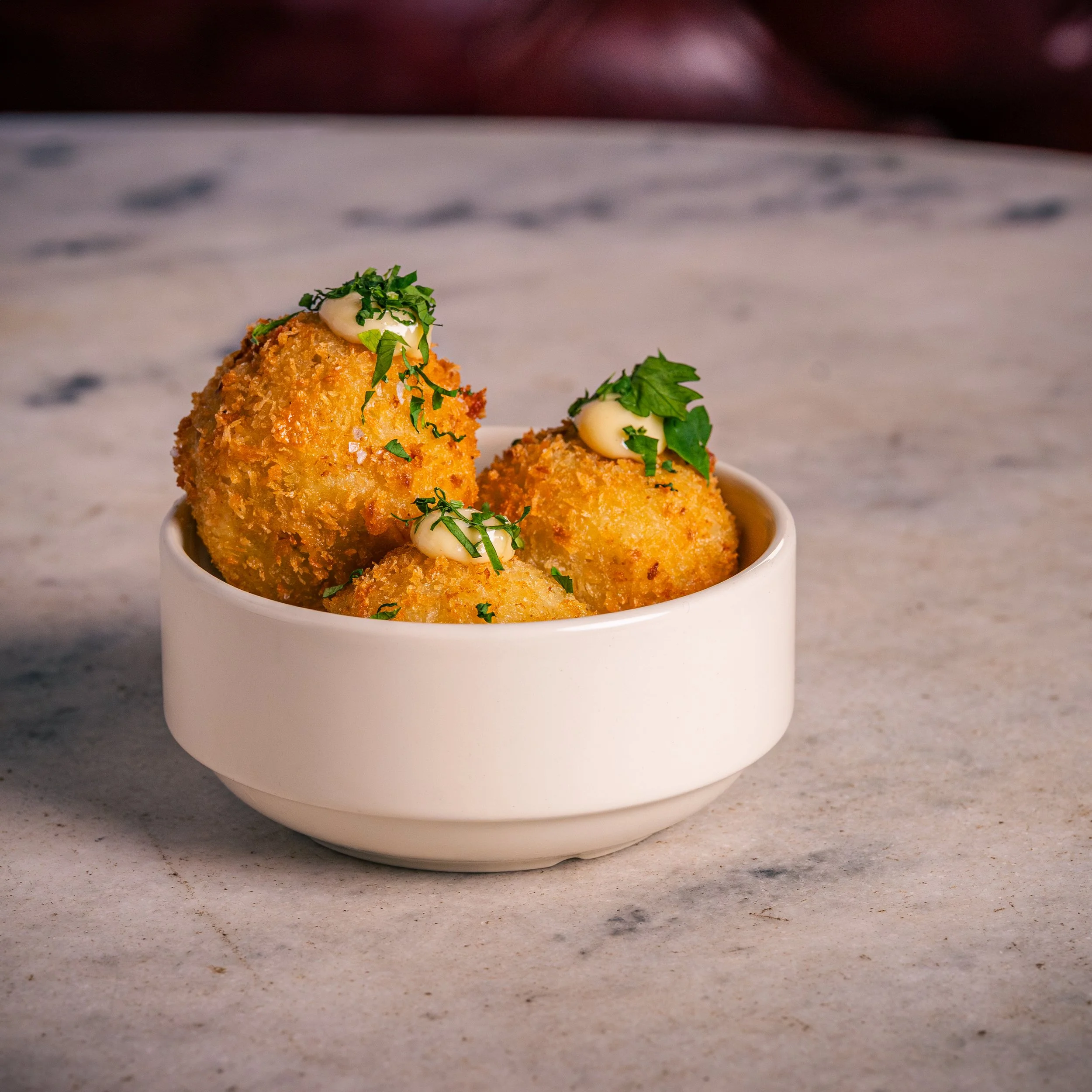 Three golden brown croquettes garnished with chopped herbs and a drizzle of white sauce in a white ceramic bowl on a marble surface.