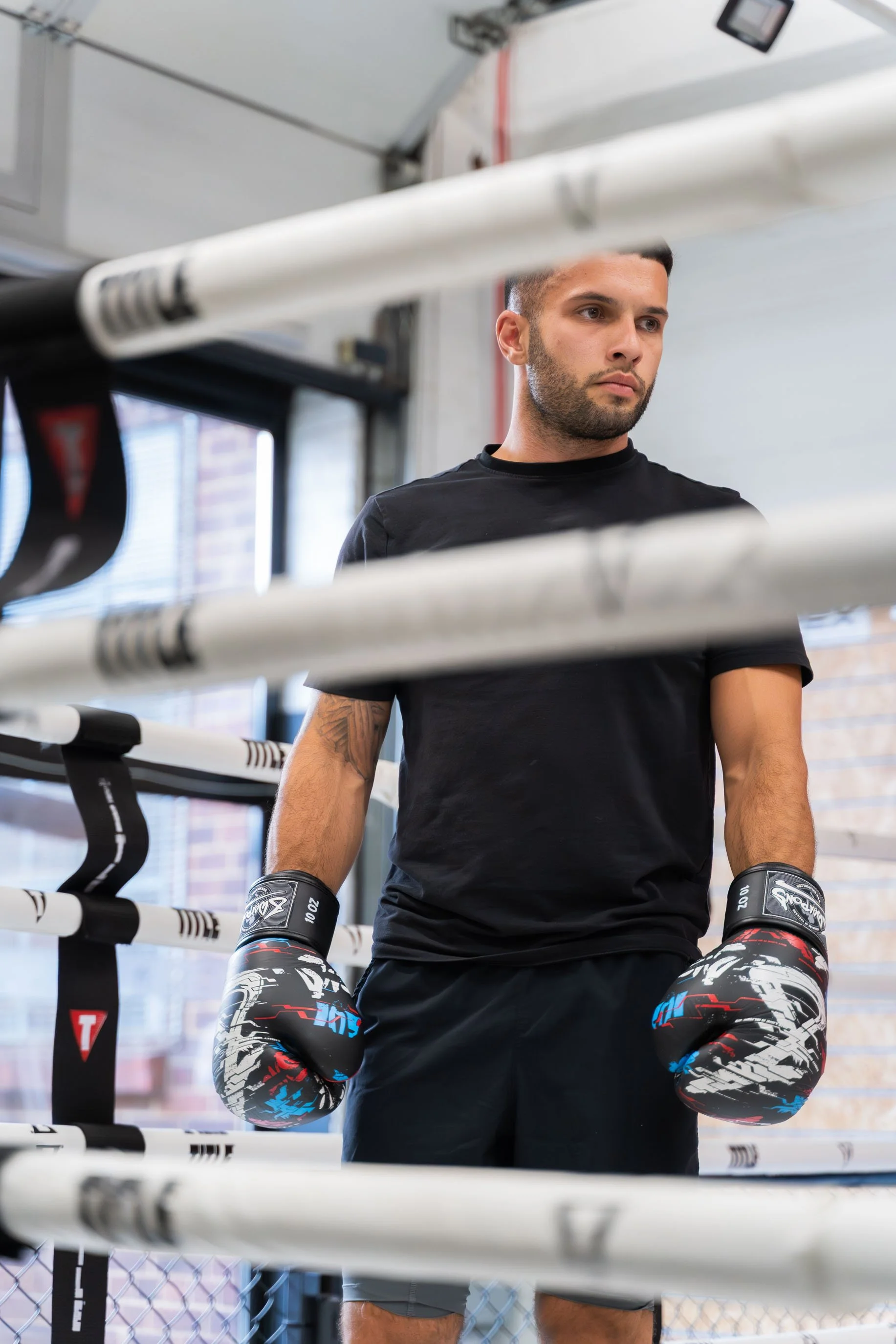 A young man with boxing gloves standing inside a boxing gym, positioned behind the ropes of a boxing ring, looking intently to the side.