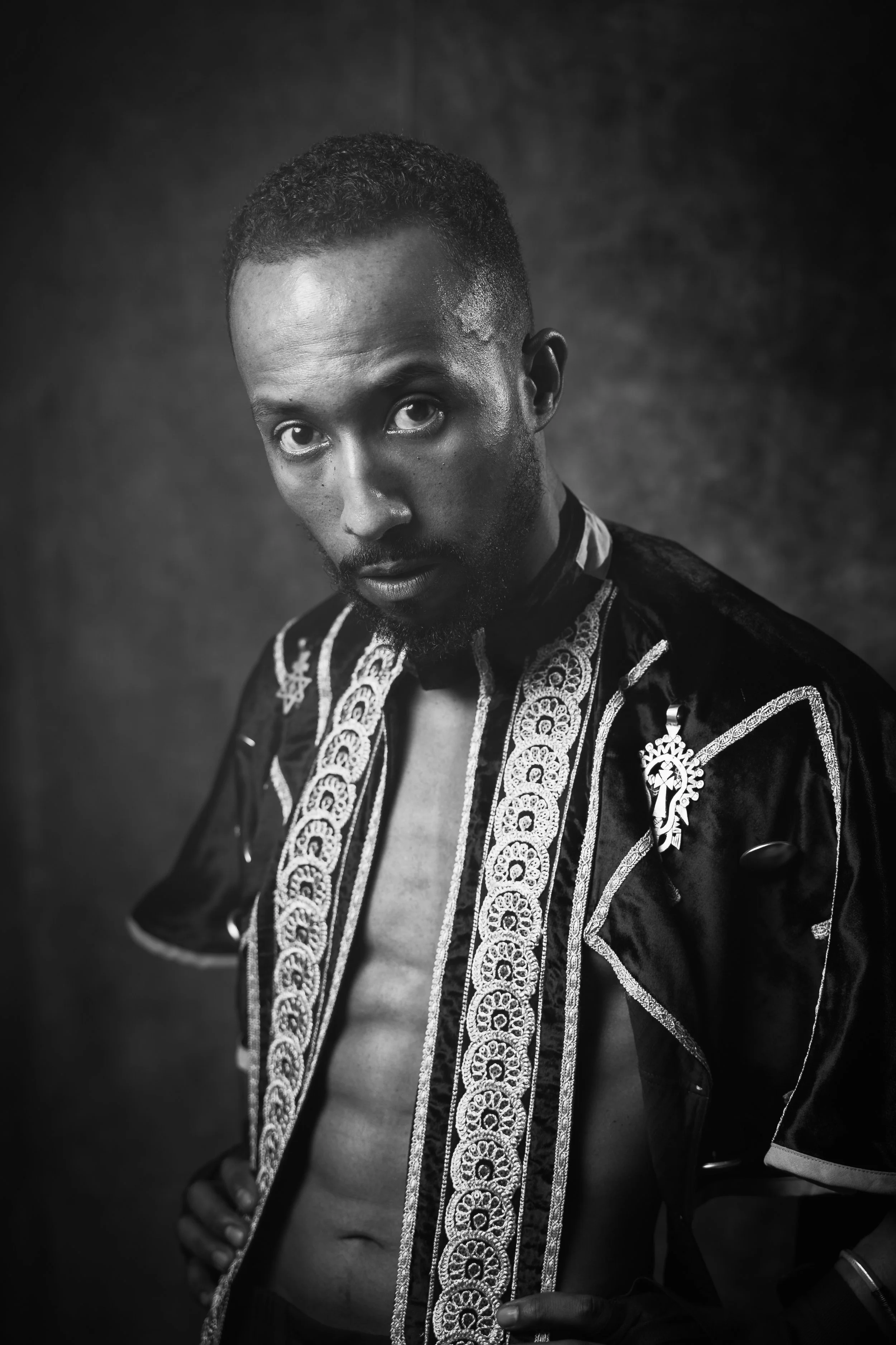 A black and white portrait of a man with short curly hair and a beard, wearing an ornate, embroidered open shirt with a partially exposed chest.