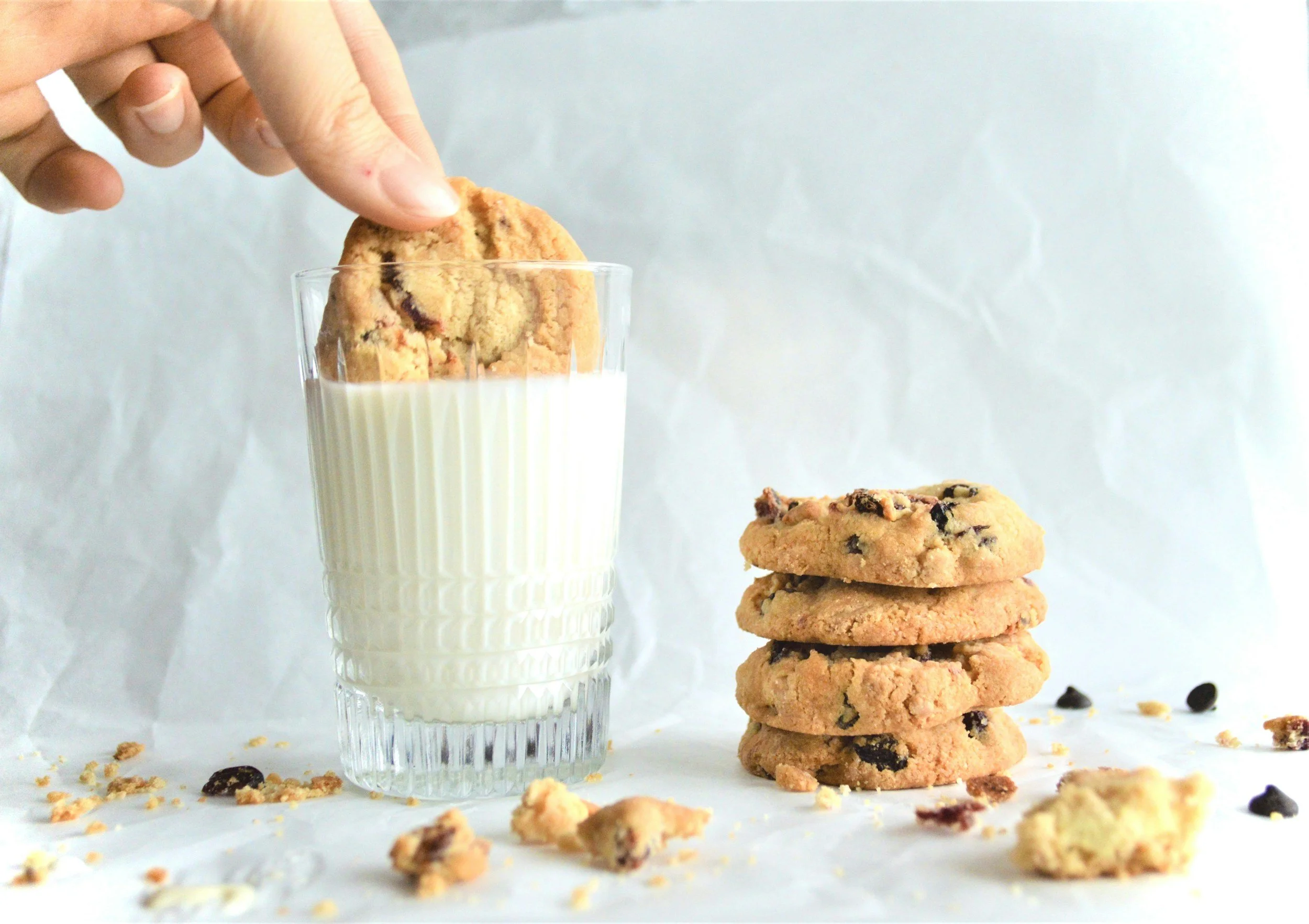 A hand dipping a chocolate chip cookie into a glass of milk, with a stack of cookies nearby and cookie crumbs scattered on a white surface.