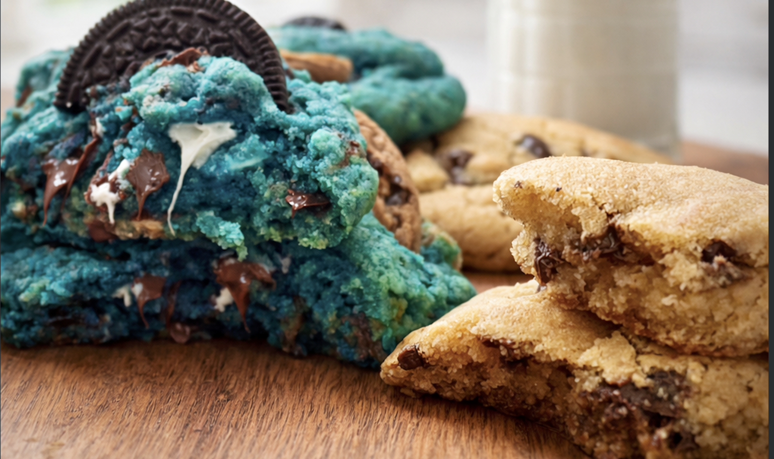 Close-up of blue and green cookies topped with an Oreo cookie and chocolate pieces, and a few chocolate chip cookies on a wooden surface.