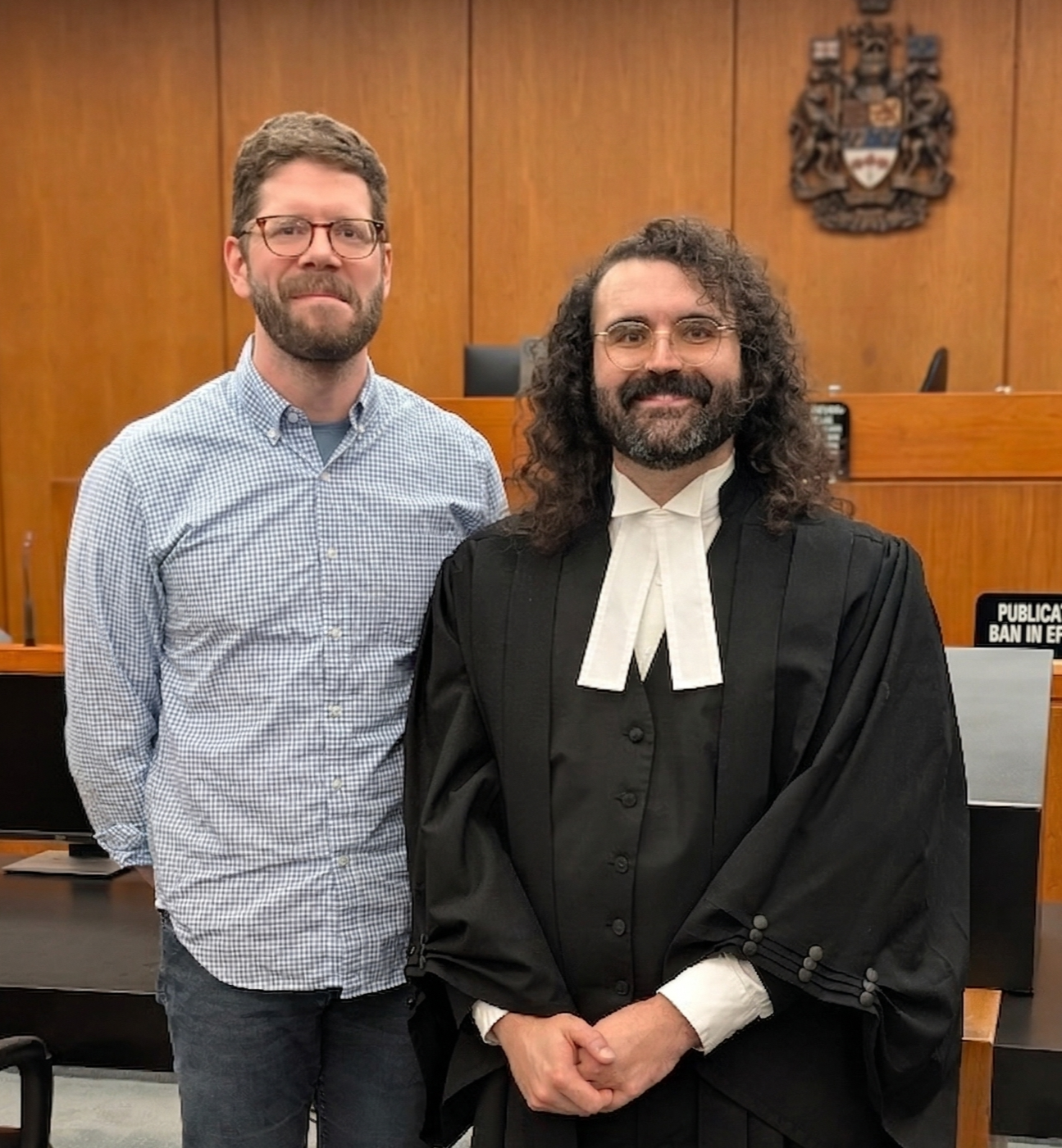 Two men, one in a checkered shirt and the other in a black judge's robe, standing inside a courtroom with wooden paneling and a judicial crest on the wall behind them.