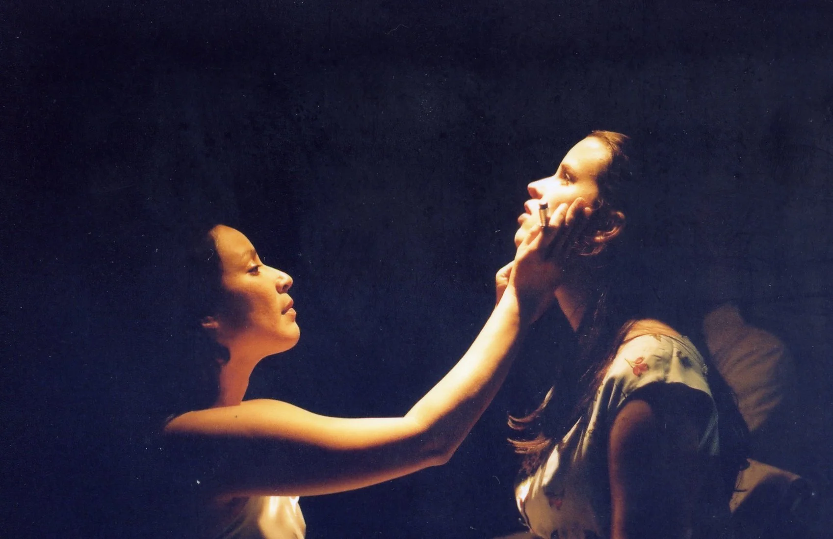 A woman is holding a young woman's face gently with her left hand, both facing each other in a dark setting with soft lighting, creating an intimate moment.