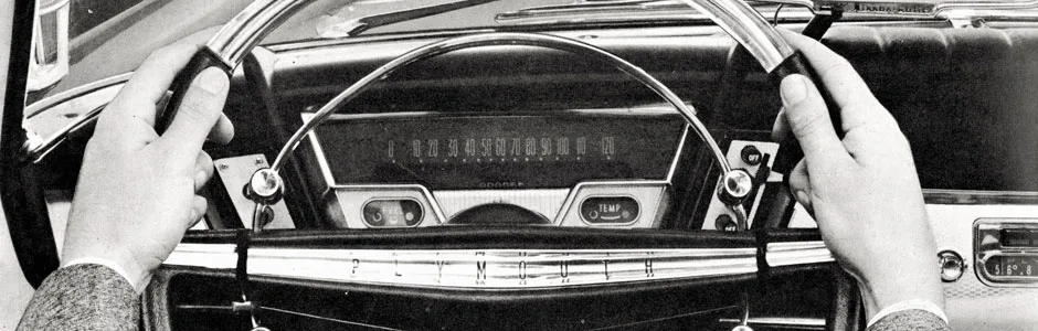 Black and white photo of a person holding a vintage steering wheel inside an old car, with a large speedometer and dashboard visible.