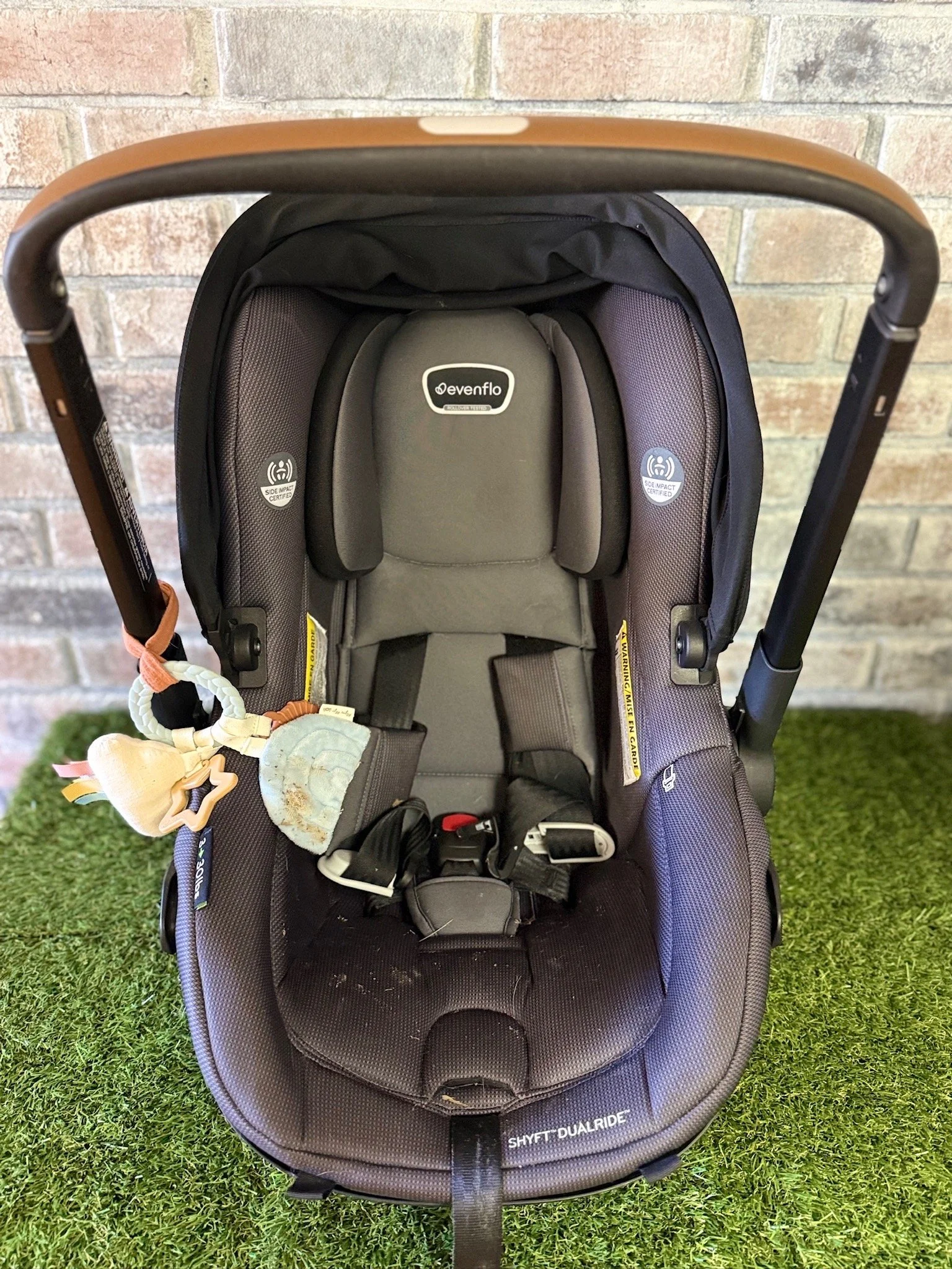 Gray infant car seat with black harness in front of a brick wall and on green grass.