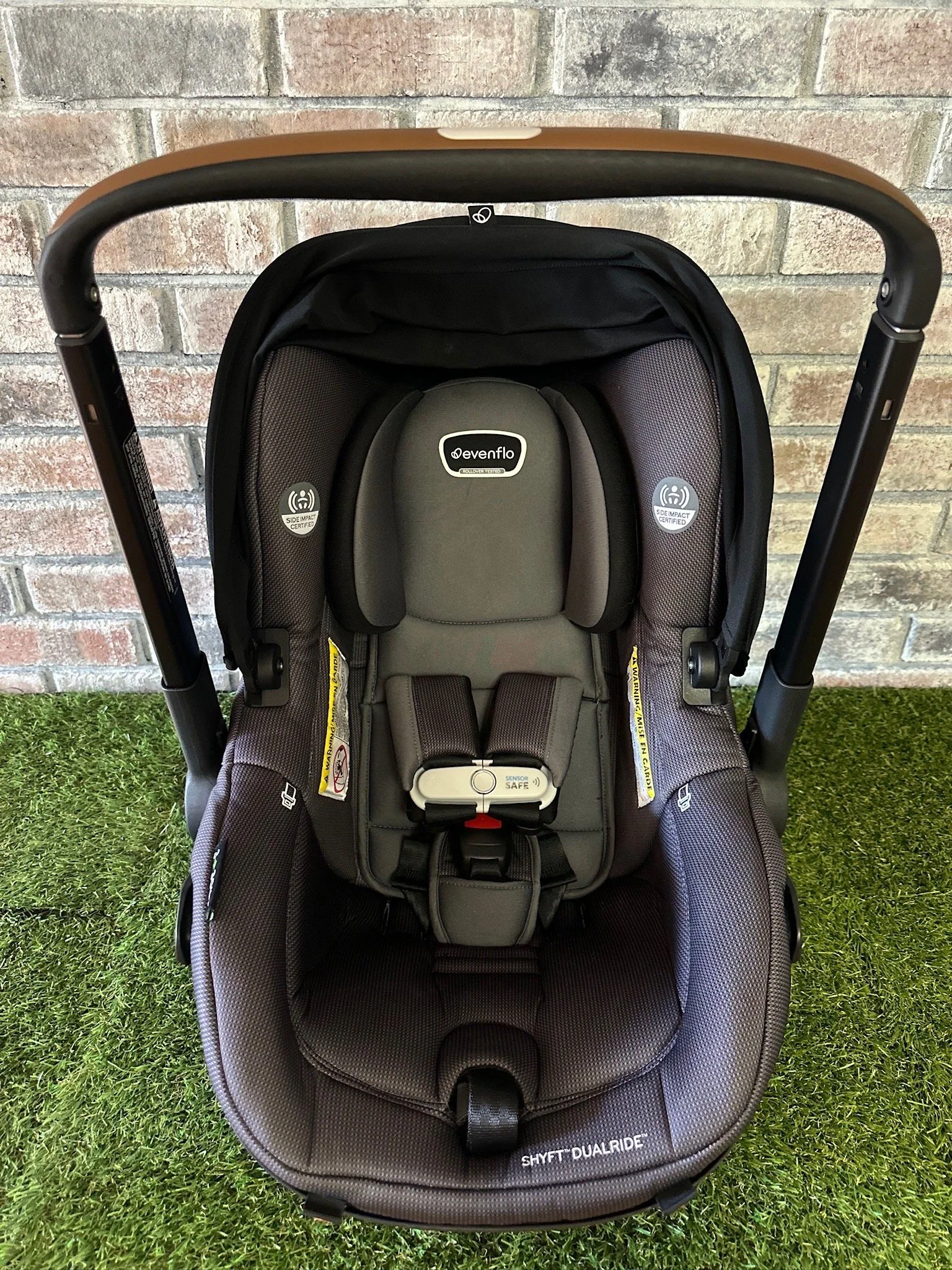 Child car seat with black and gray fabric, labeled 'SHYFT DUALRIDE', placed on grass against a brick wall.