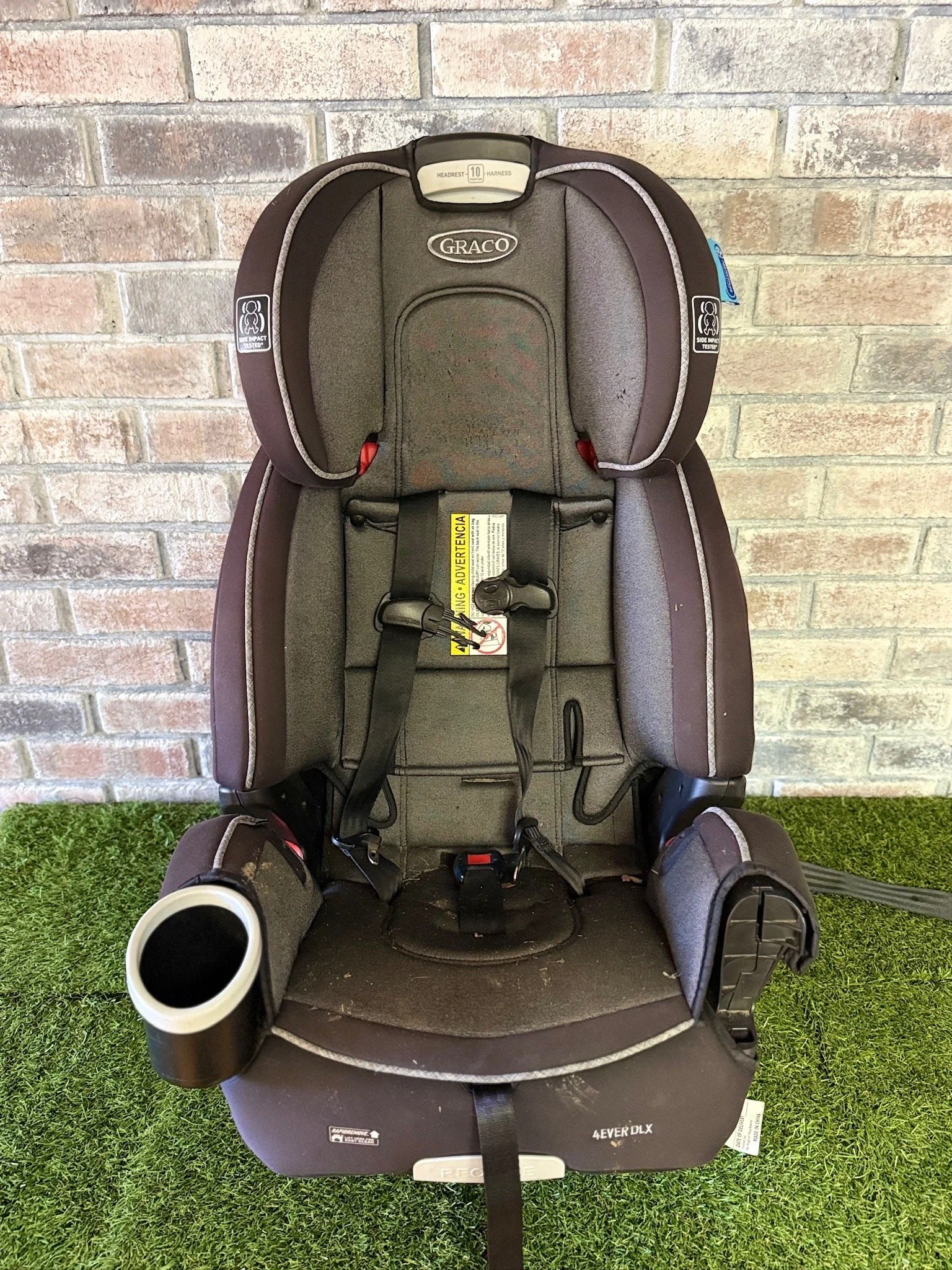 Child car seat with head support, harness, and cup holder, placed in front of a brick wall on artificial grass.