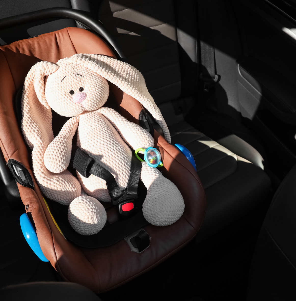 A stuffed bunny toy with long ears and a pink nose sits in a child's car seat on the back seat of a vehicle. The car seat is brown with black straps.