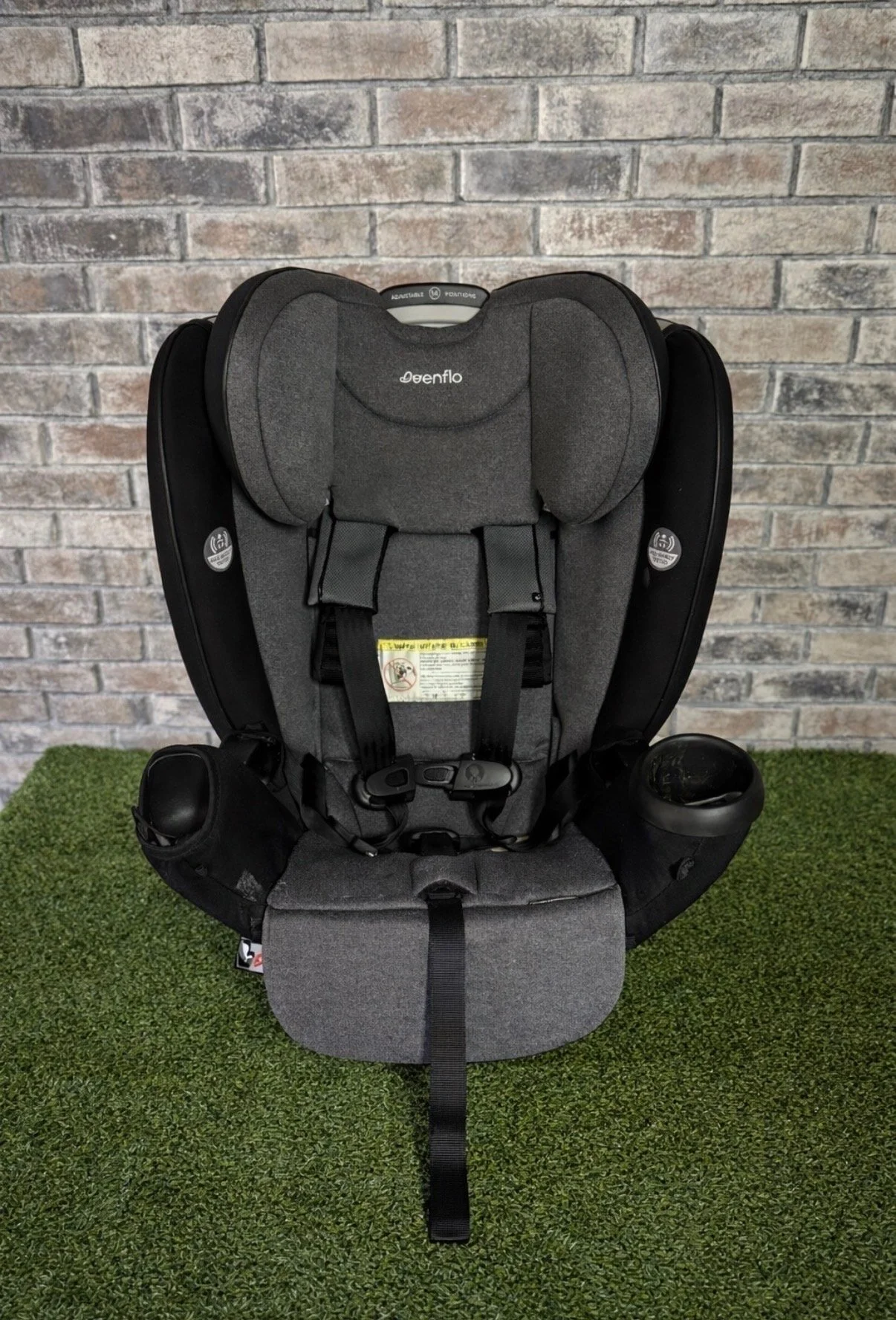 Gray child car seat with black straps, head support, and side padding, positioned on green artificial grass in front of a brick wall.
