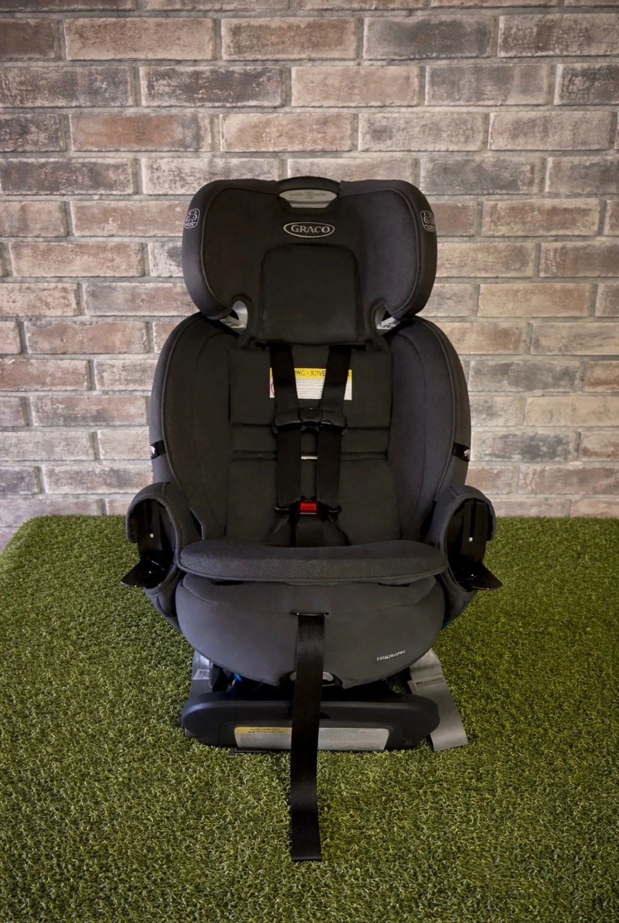 Black child car seat with headrest and harness, placed on green textured surface against a brick wall background.
