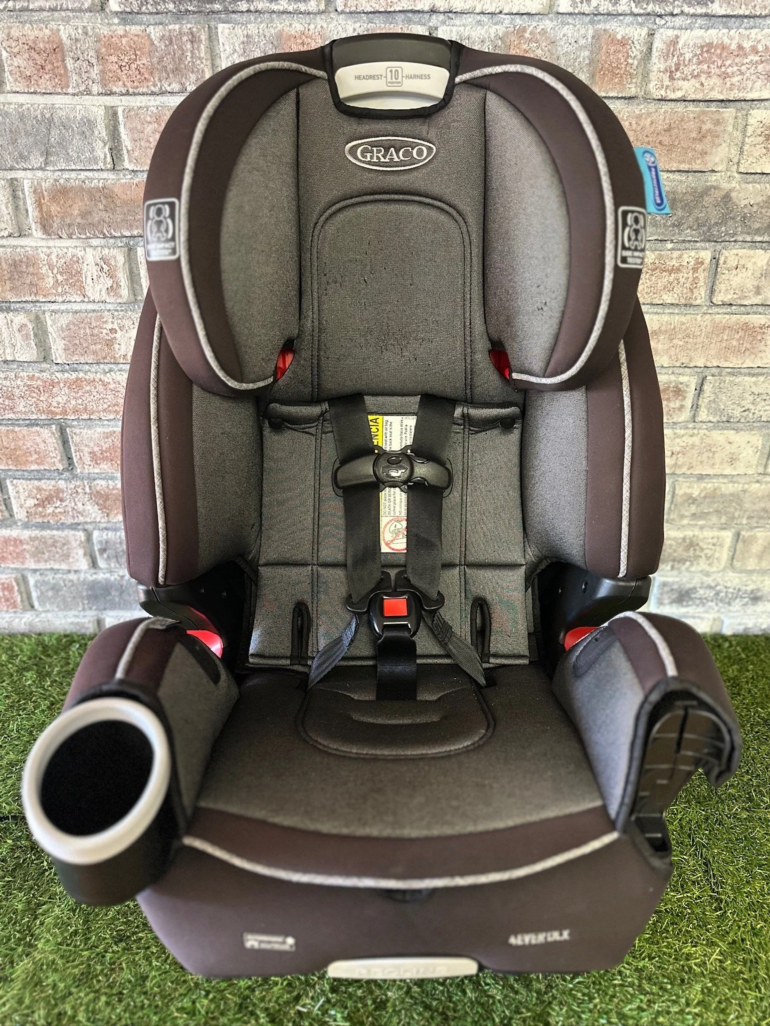 Gray and black Graco child car seat with headrest, shoulder straps, and harness, placed on green artificial grass in front of a brick wall.