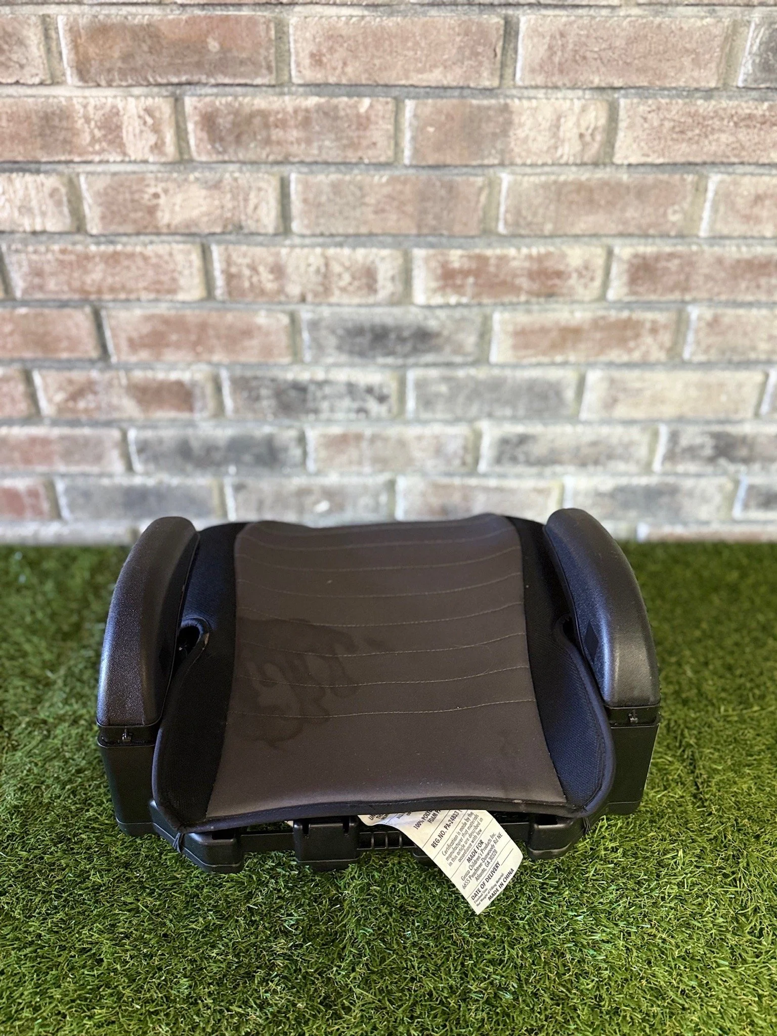 A black child car seat with side supports, placed on green artificial turf, against a brick wall background.