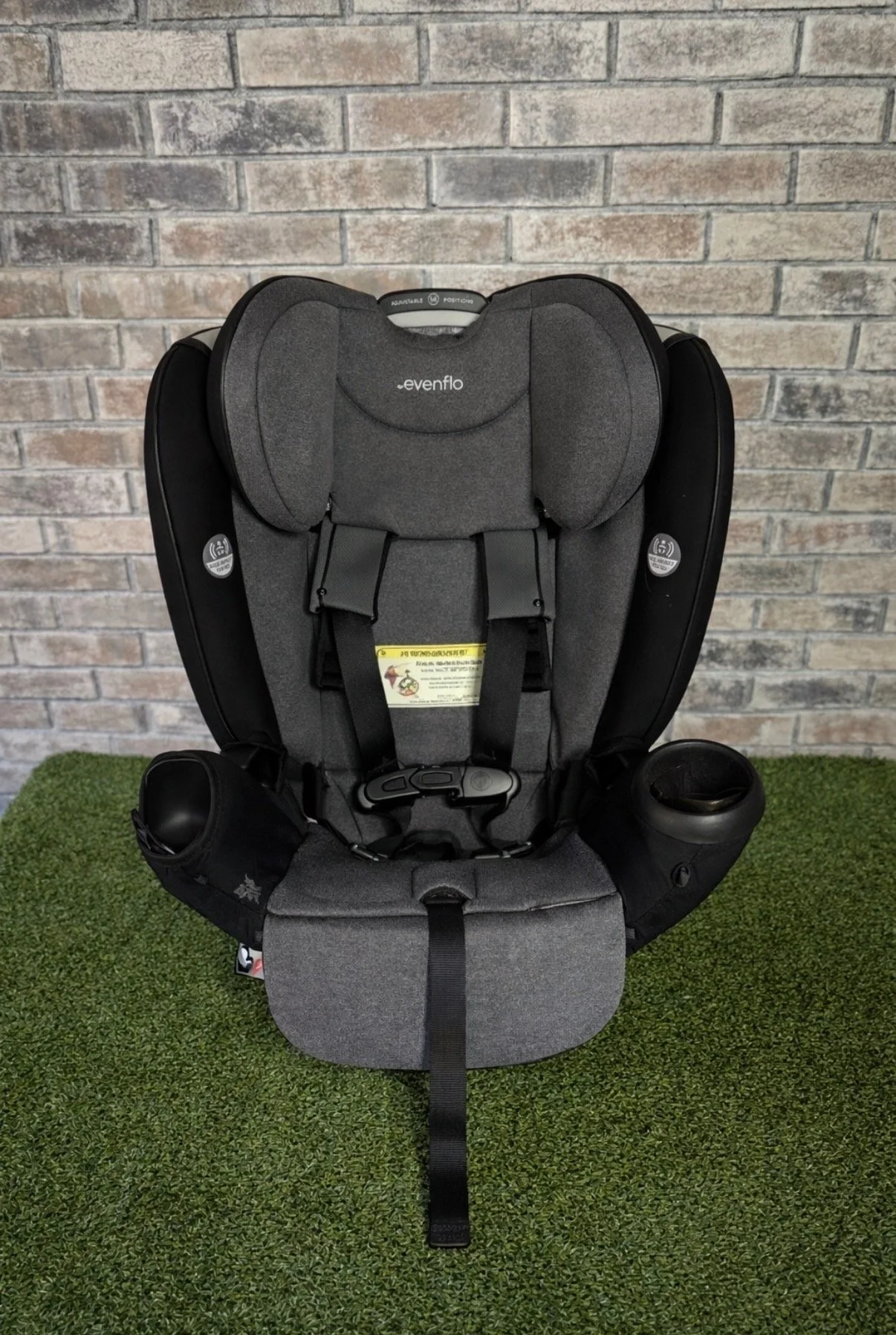 A black and gray child car seat with adjustable head support, shoulder straps, and cup holders on each side, placed on green artificial grass against a brick wall background.