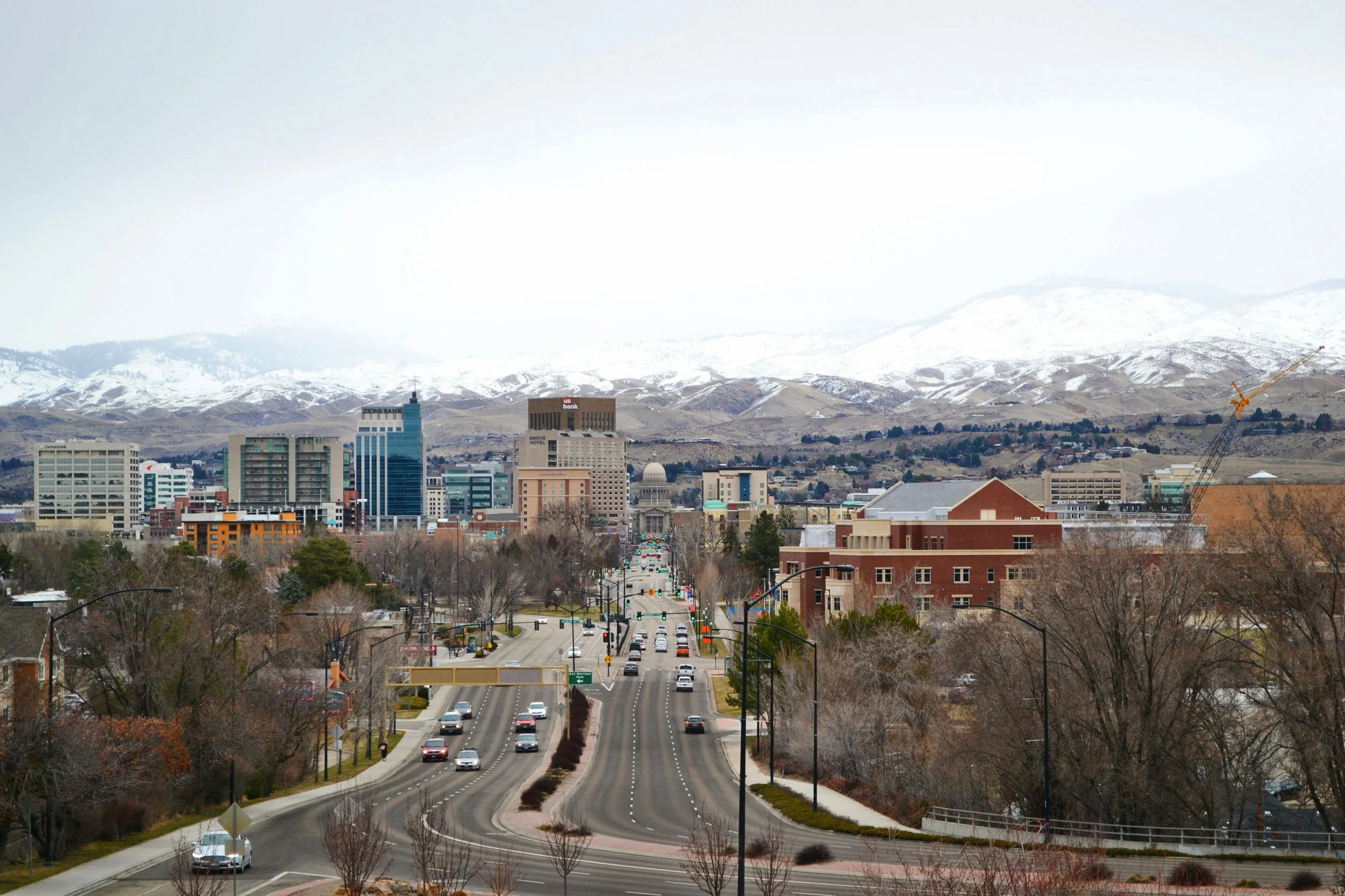 Boise Idaho road scene with cars, buildings, leafless trees, and snow-capped mountains in the background.
