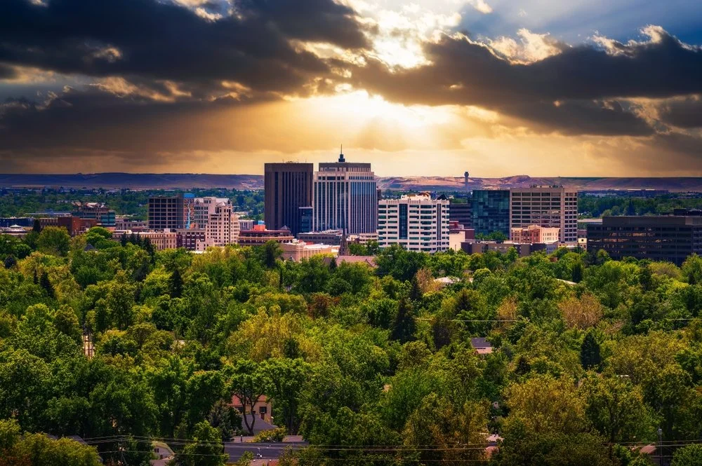 Boise, Idaho skyline with tall buildings and lush green trees in the foreground, during sunset with clouds and sunlight breaking through.