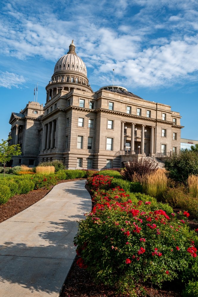 The image shows the state capitol building in Boise, Idaho with a prominent dome on top, surrounded by well-maintained gardens and a curved walkway leading to the entrance.