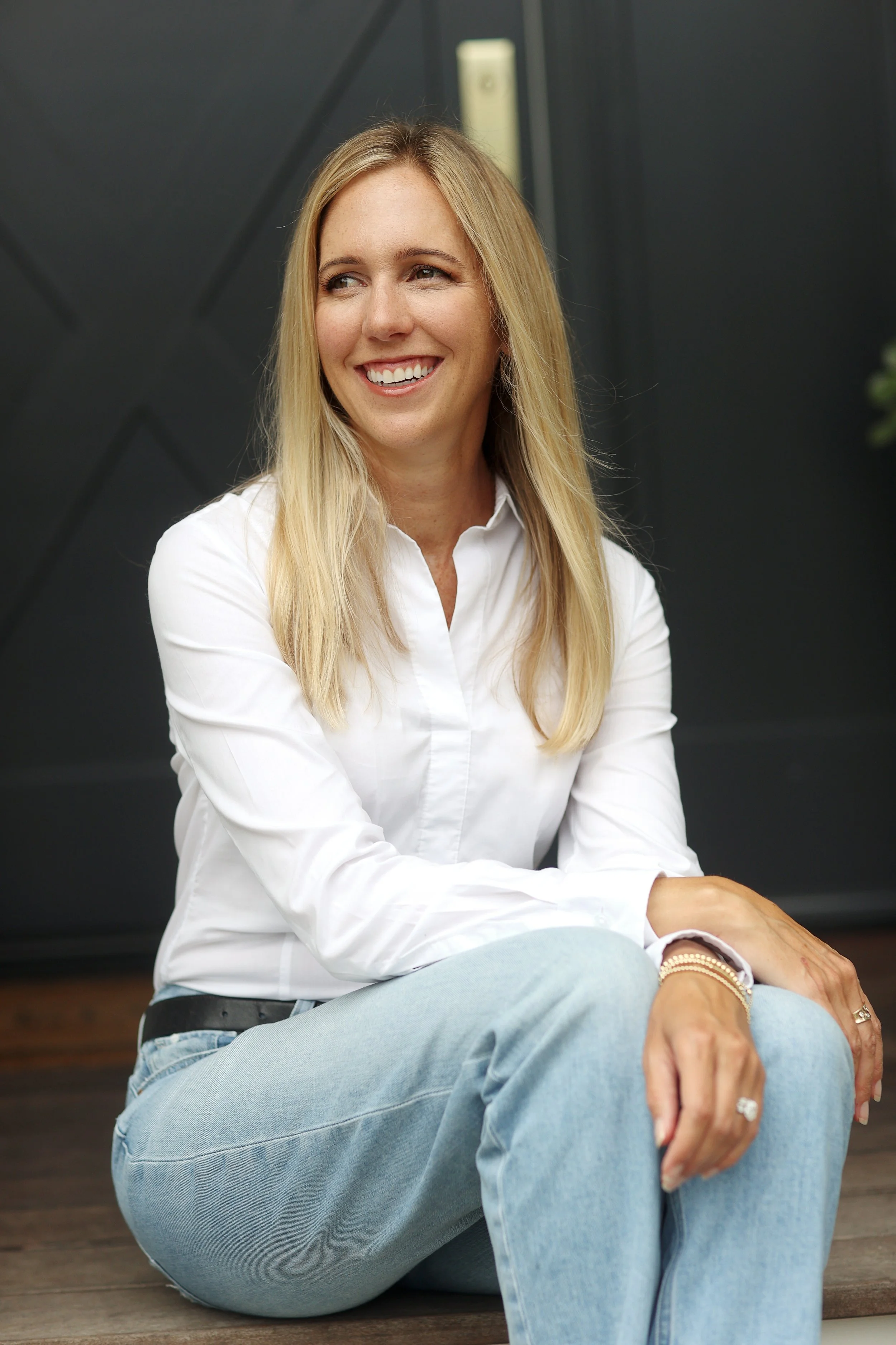 A woman with long blonde hair, wearing a white shirt and light blue jeans, sitting on wooden steps, smiling and looking to the side.