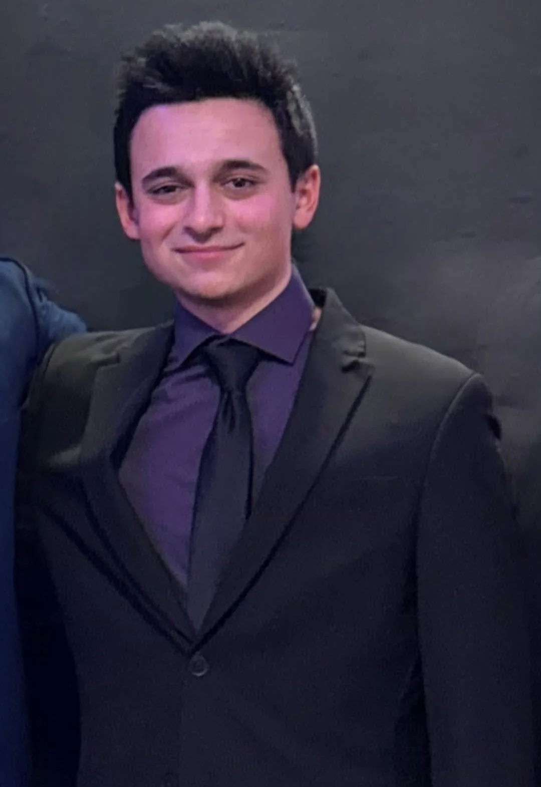 A young man with dark hair, wearing a black suit, purple shirt, and black tie, smiling against a dark background.