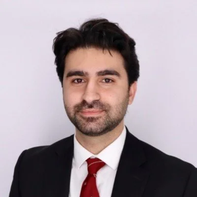A man in a black suit, white shirt, and red tie, with dark hair and a beard, smiling at the camera.