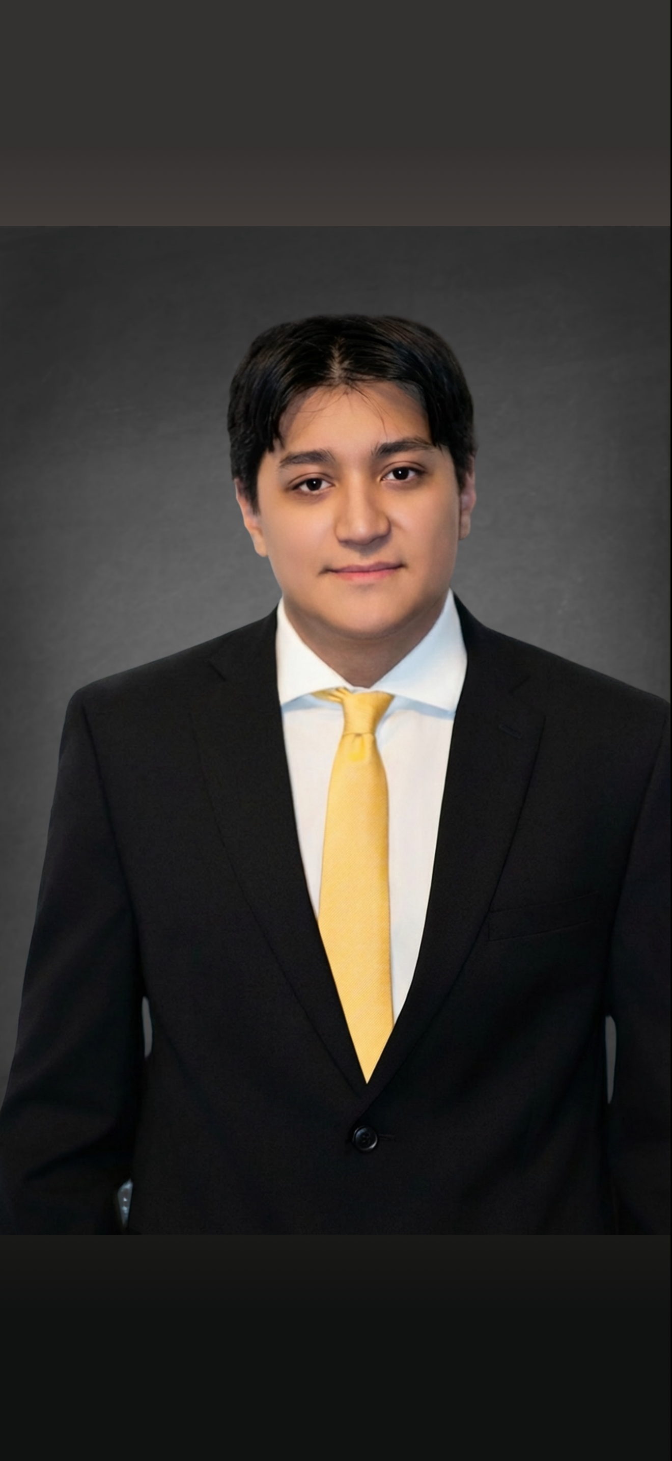Young man in formal business attire wearing a black suit, white shirt, and yellow tie, posing against a gray background.