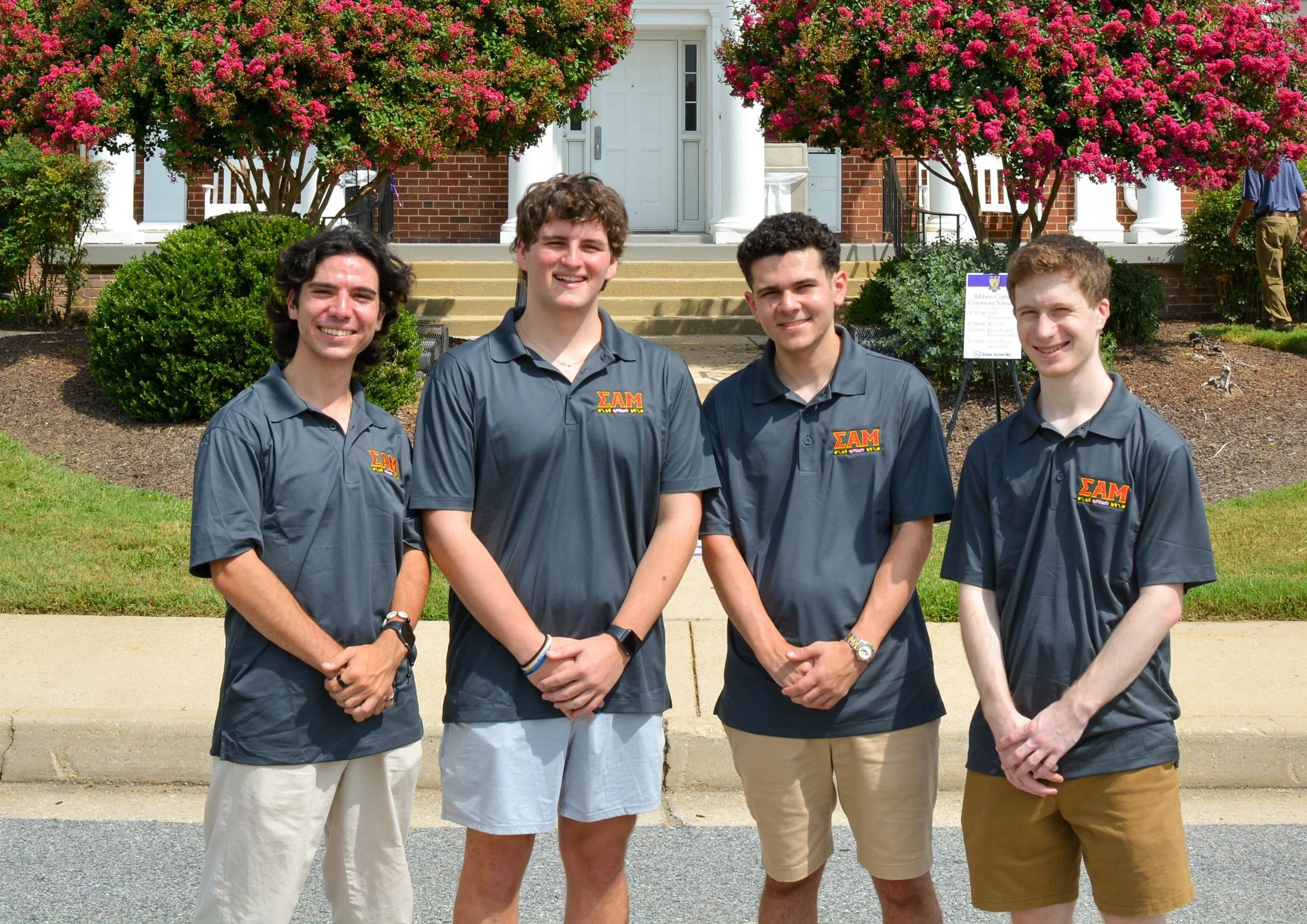 Four young men standing together outdoors in front of a house with flowering bushes. They are smiling and wearing dark shirts with a Greek letters logo. Two are wearing khaki shorts and one is wearing white shorts.