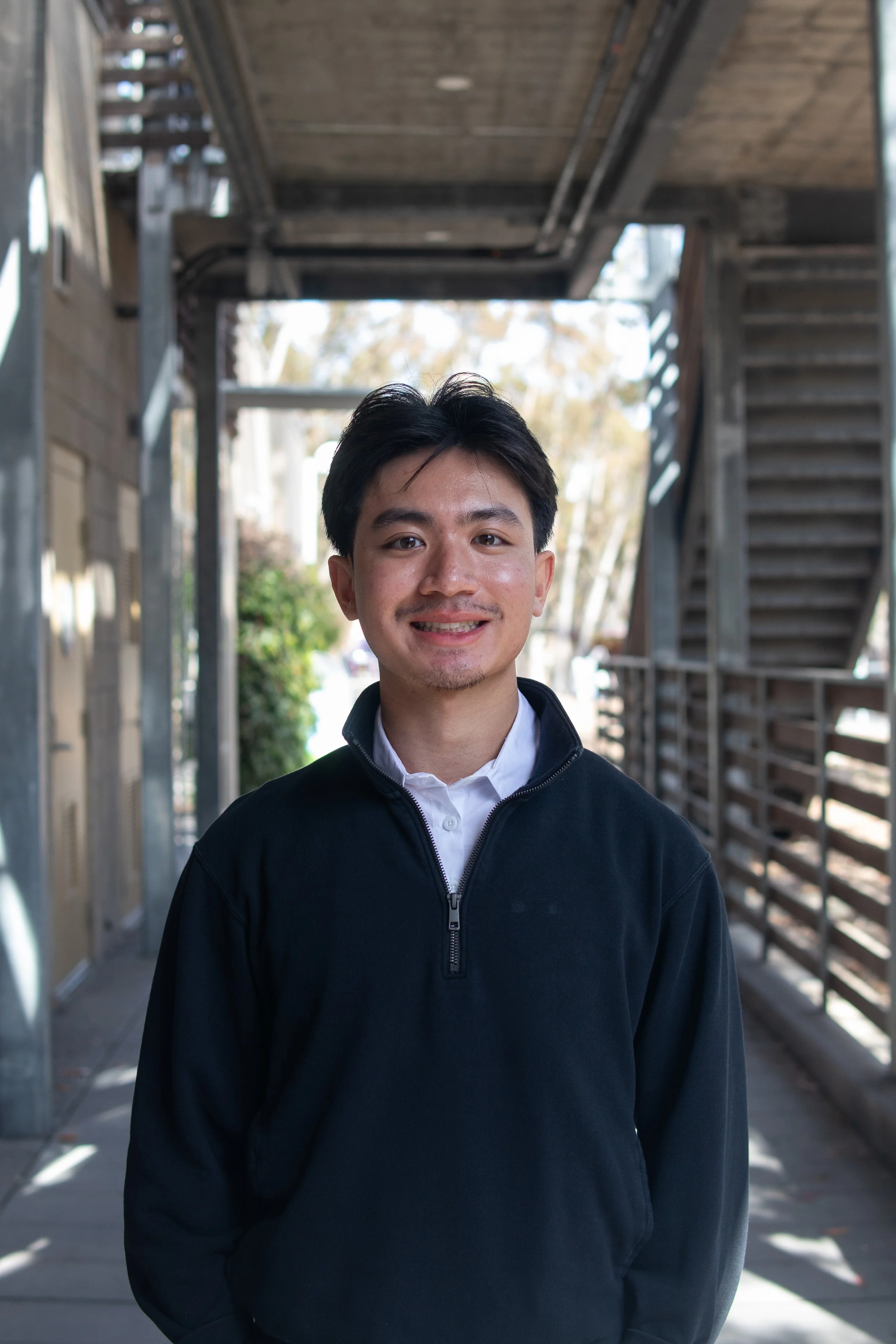 A young man standing outdoors under a wooden walkway with stairs, wearing a black zip-up jacket and smiling.