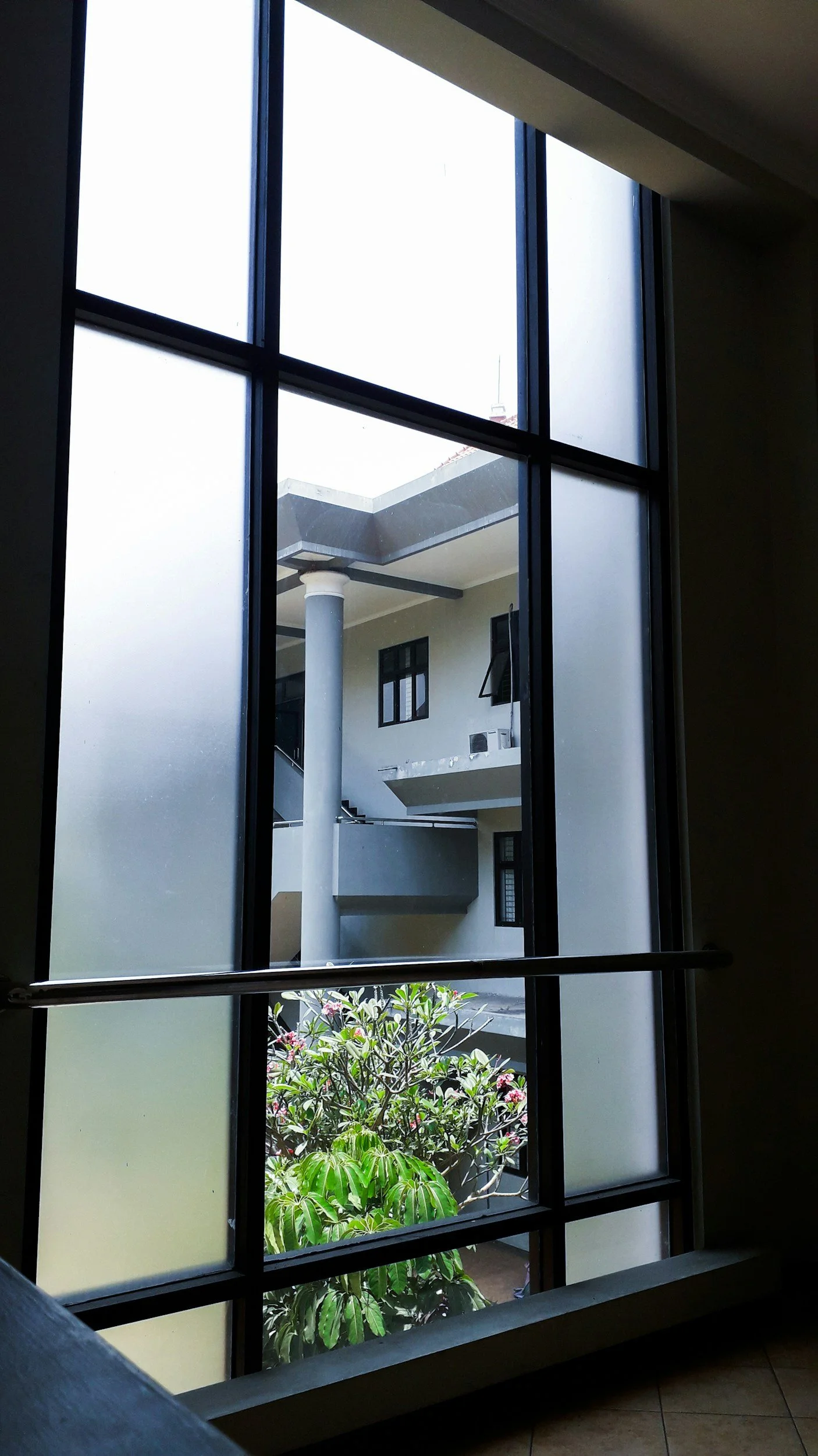 View of an apartment complex seen through a window with frosted and clear glass panels, black window frame, green plants outside, building with balconies and windows.