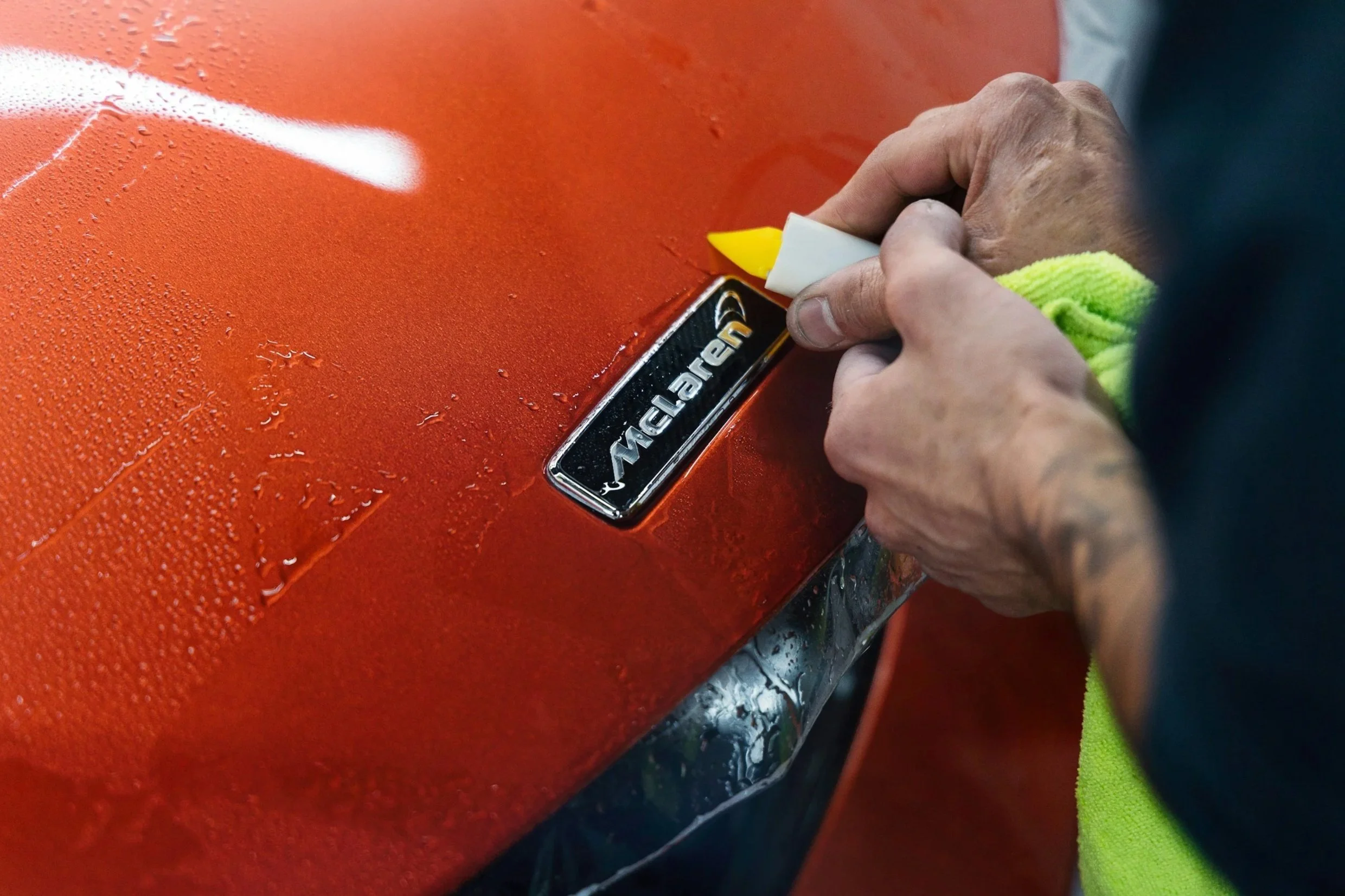 Professional installer applying a paint protection film around the badging of  a red vehicle with water droplets on the surface.